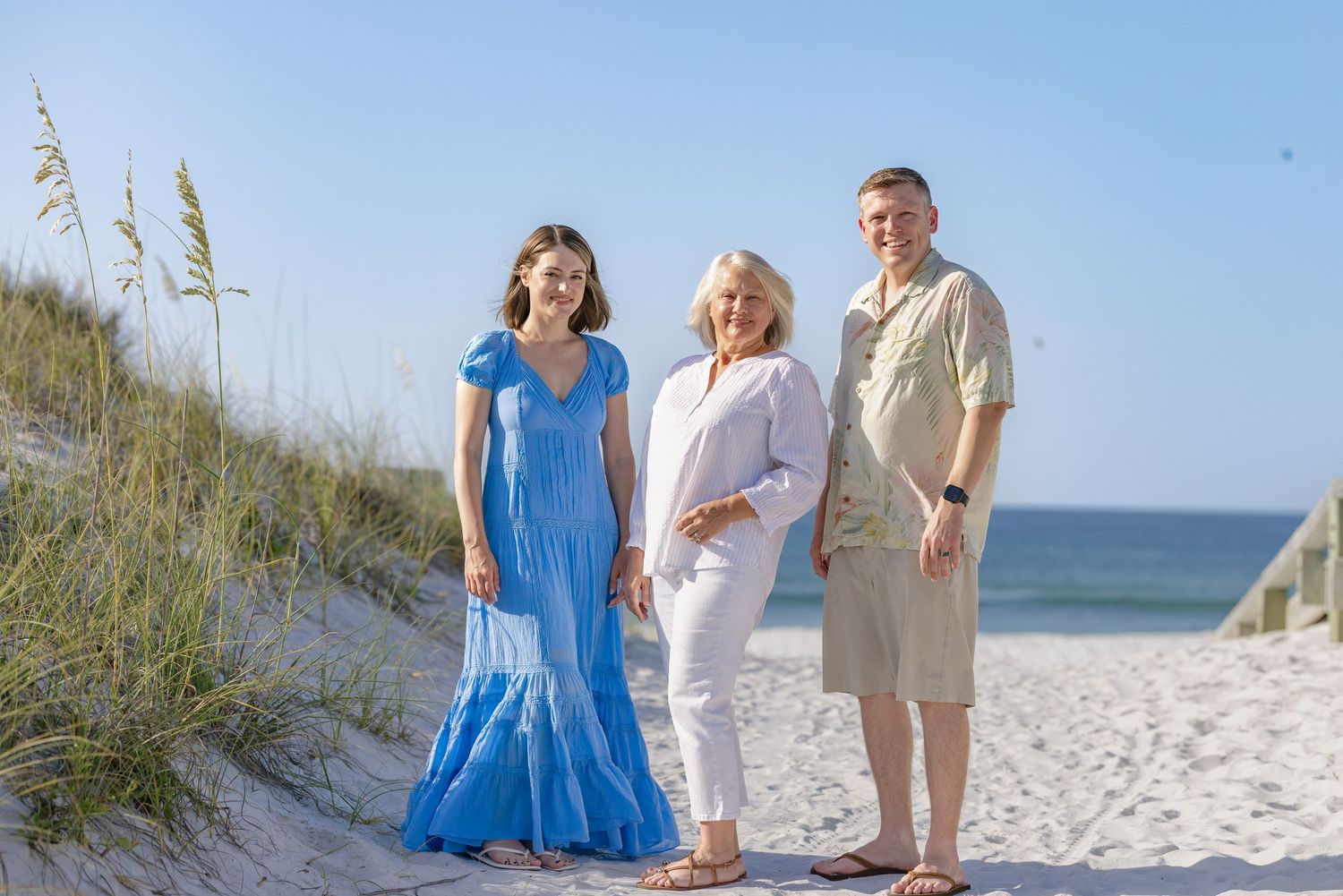 Family of three posing on sandy beach path with sea oats, ocean in background, sunny day.