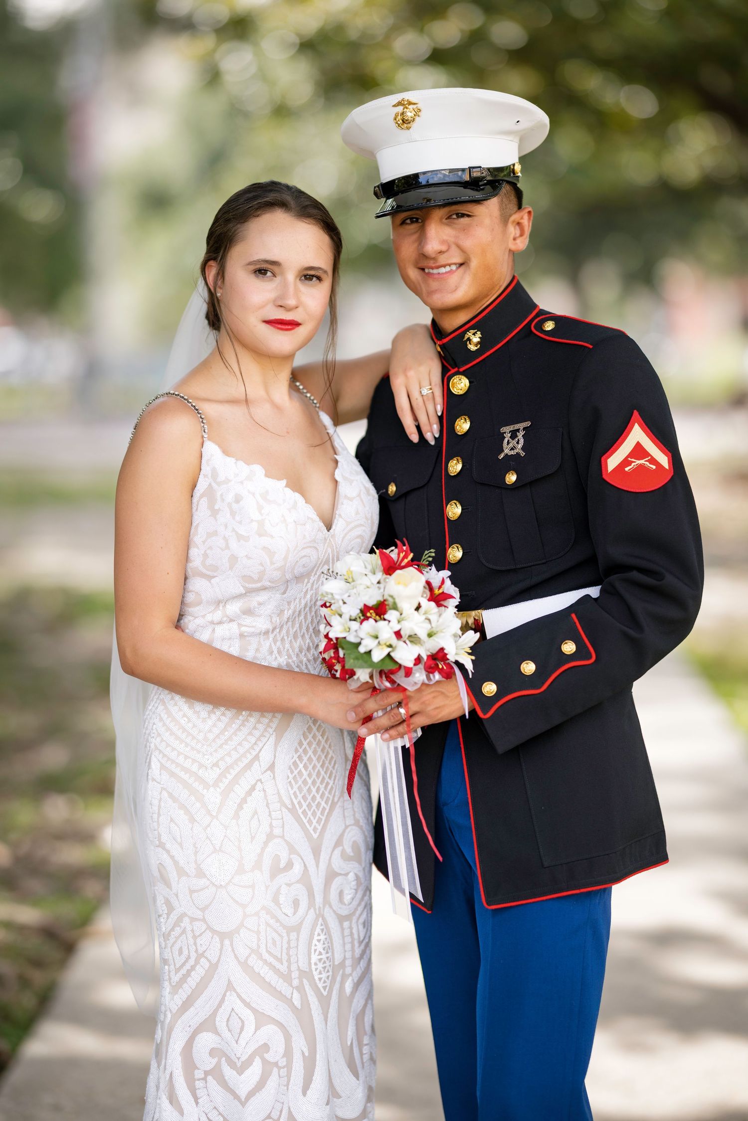 Marine in dress blues and bride in white lace gown pose together holding red and white bouquet on wedding day.