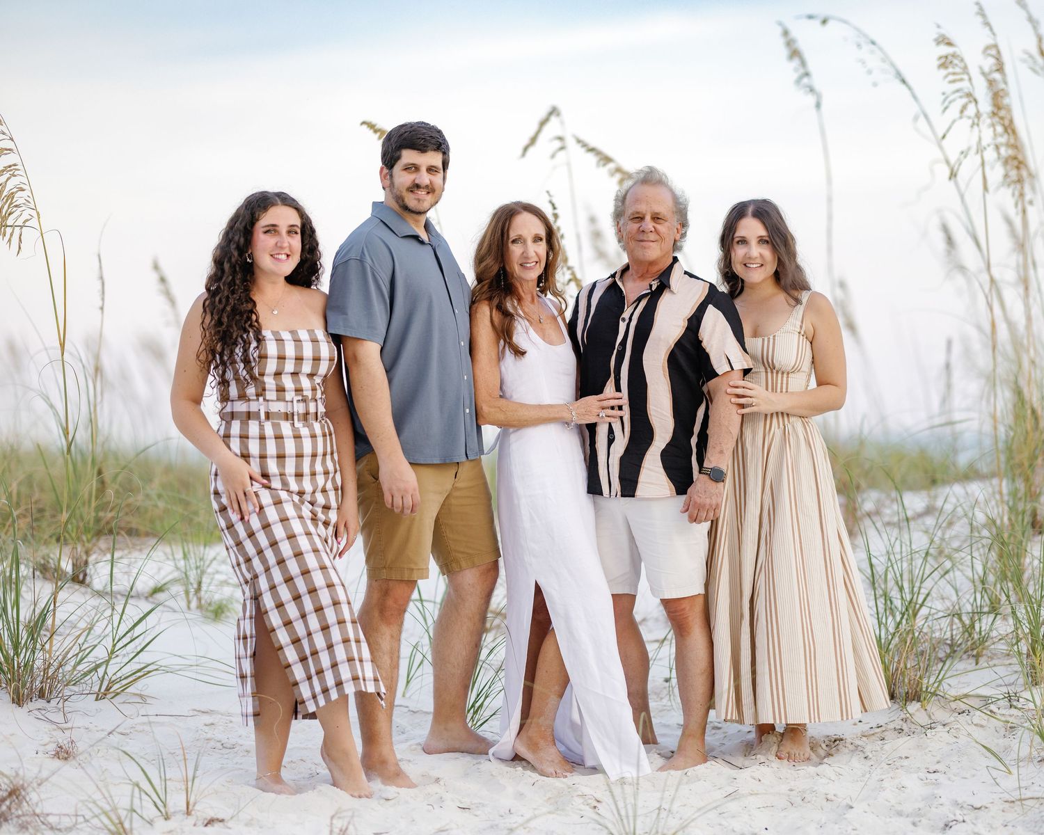Family of five posing together on a sandy beach with sea oats, dressed in coordinated outfits for a portrait.