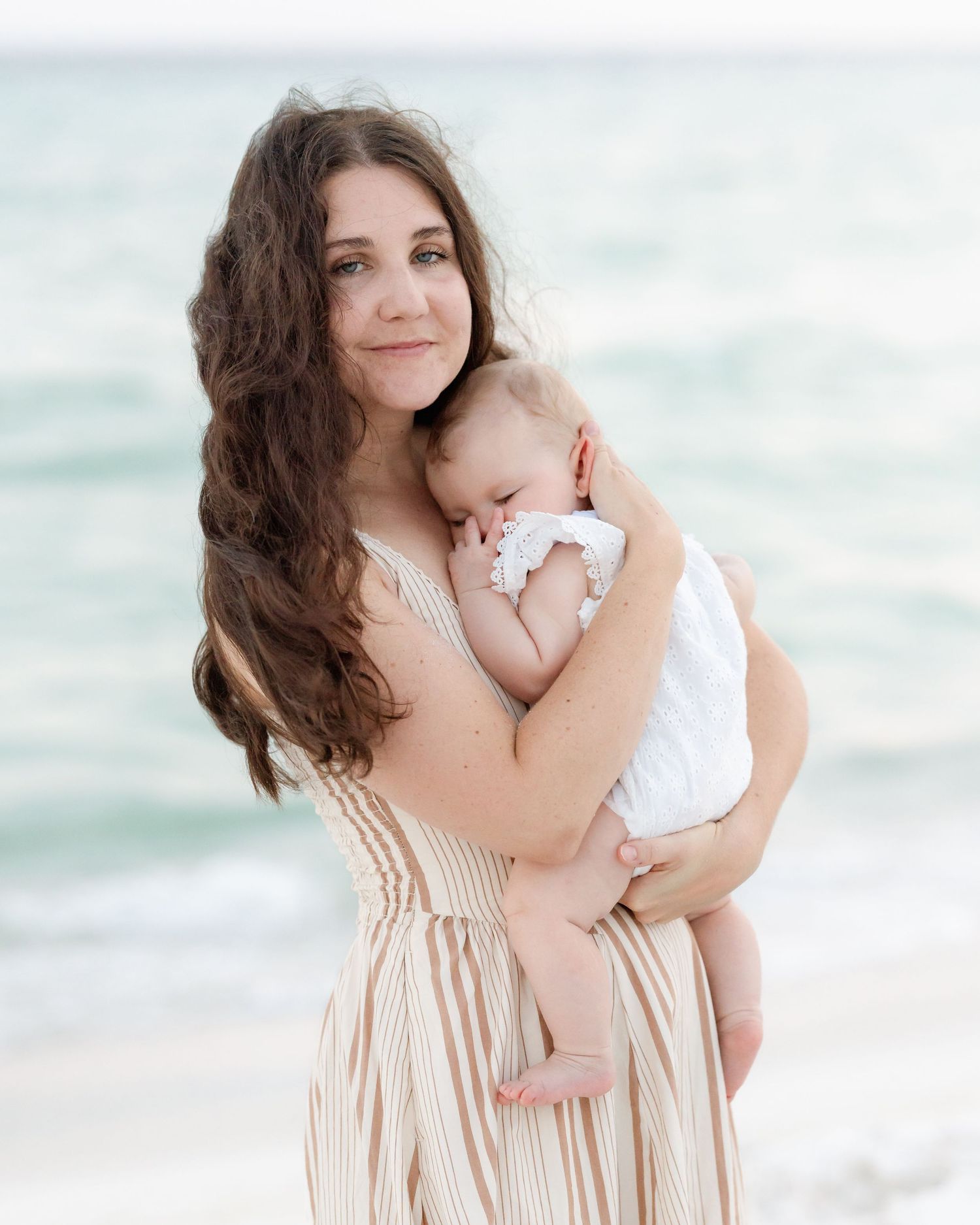 Mother in striped dress holds sleeping baby at beach.