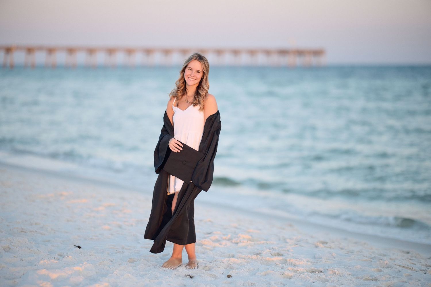 A person in a black dress walks along a white sandy beach at sunset with ocean waves in the background.