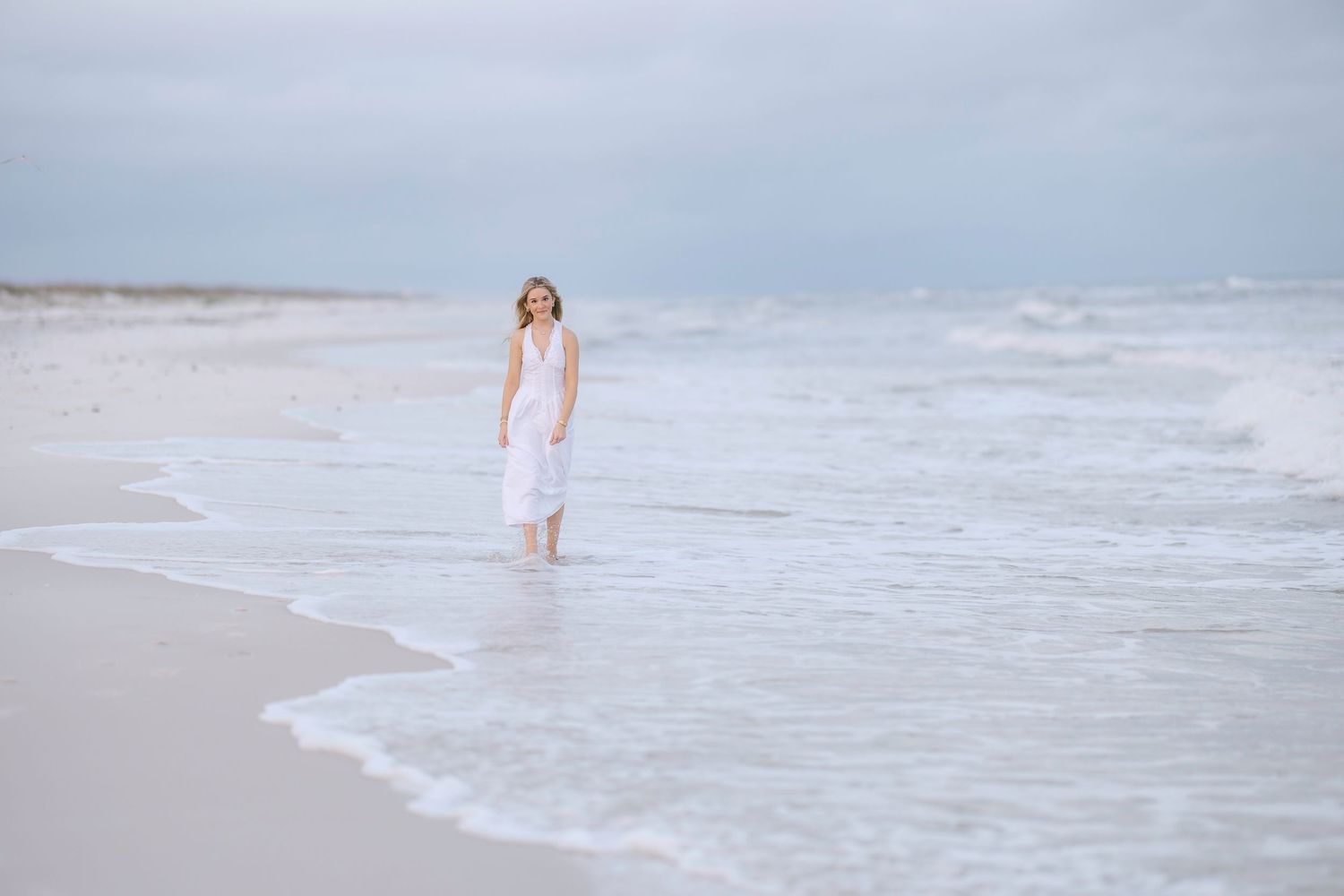 A person in white clothing walks along a misty beach shoreline as gentle waves roll in at sunset.