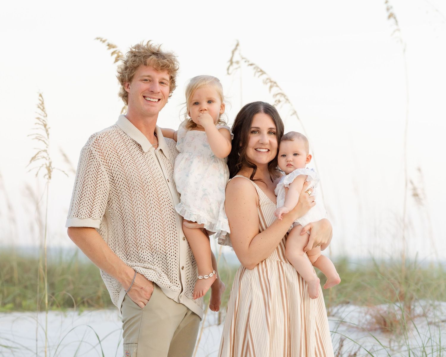 Happy family of four posing on a beach with sea oats, parents holding two young children in soft natural light.