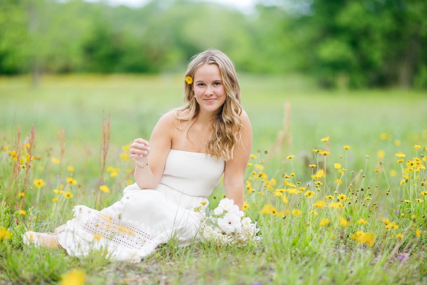 Young blonde woman in white strapless dress sitting in yellow wildflower field with flower in hair.