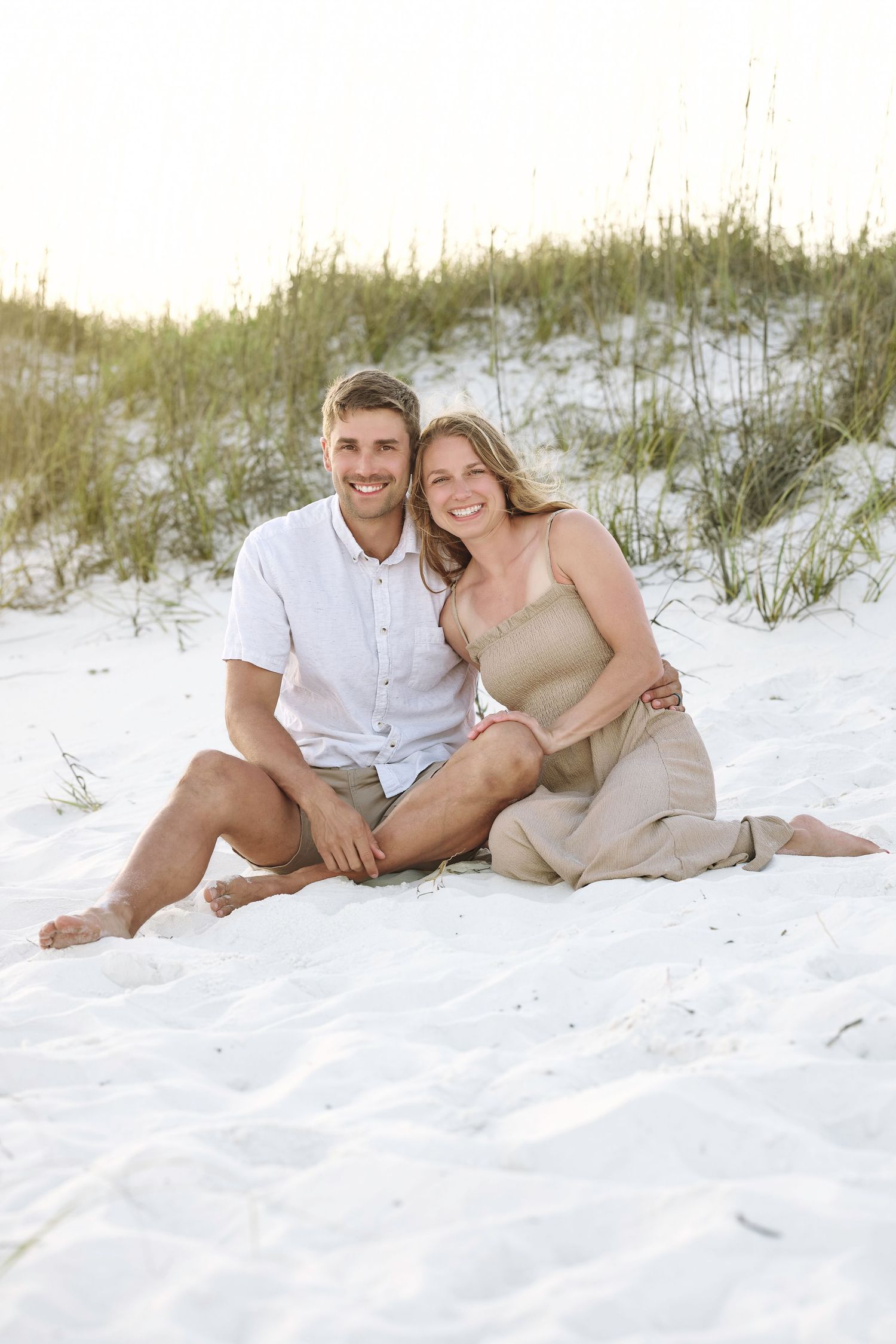 Happy couple sitting together on white sand beach dunes, smiling, wearing casual summer outfits.