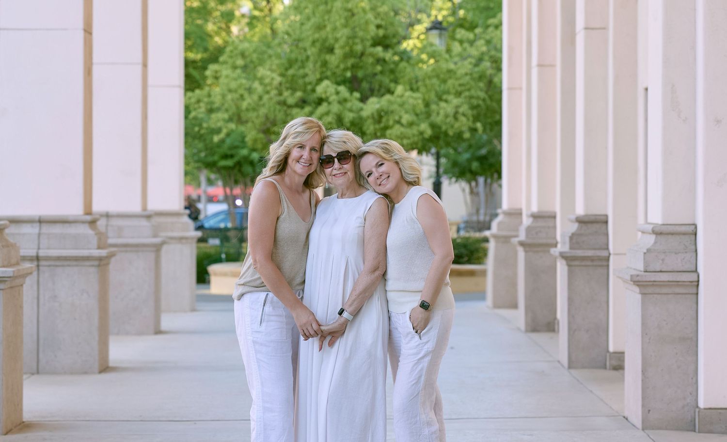 Three people in white outfits stand together in an elegant columned walkway with greenery visible in background.