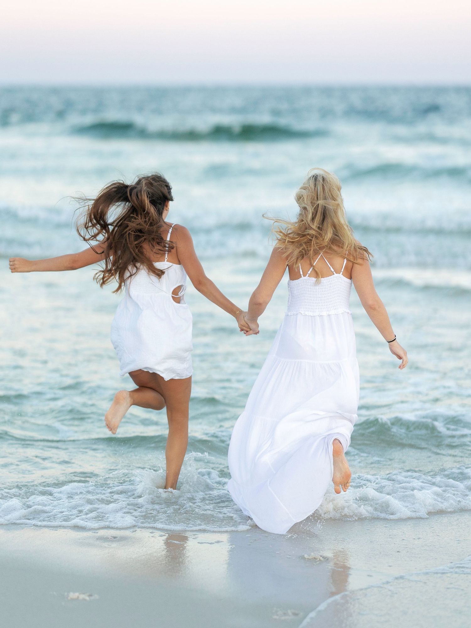 Two friends in white dresses running joyfully through ocean waves at sunset.