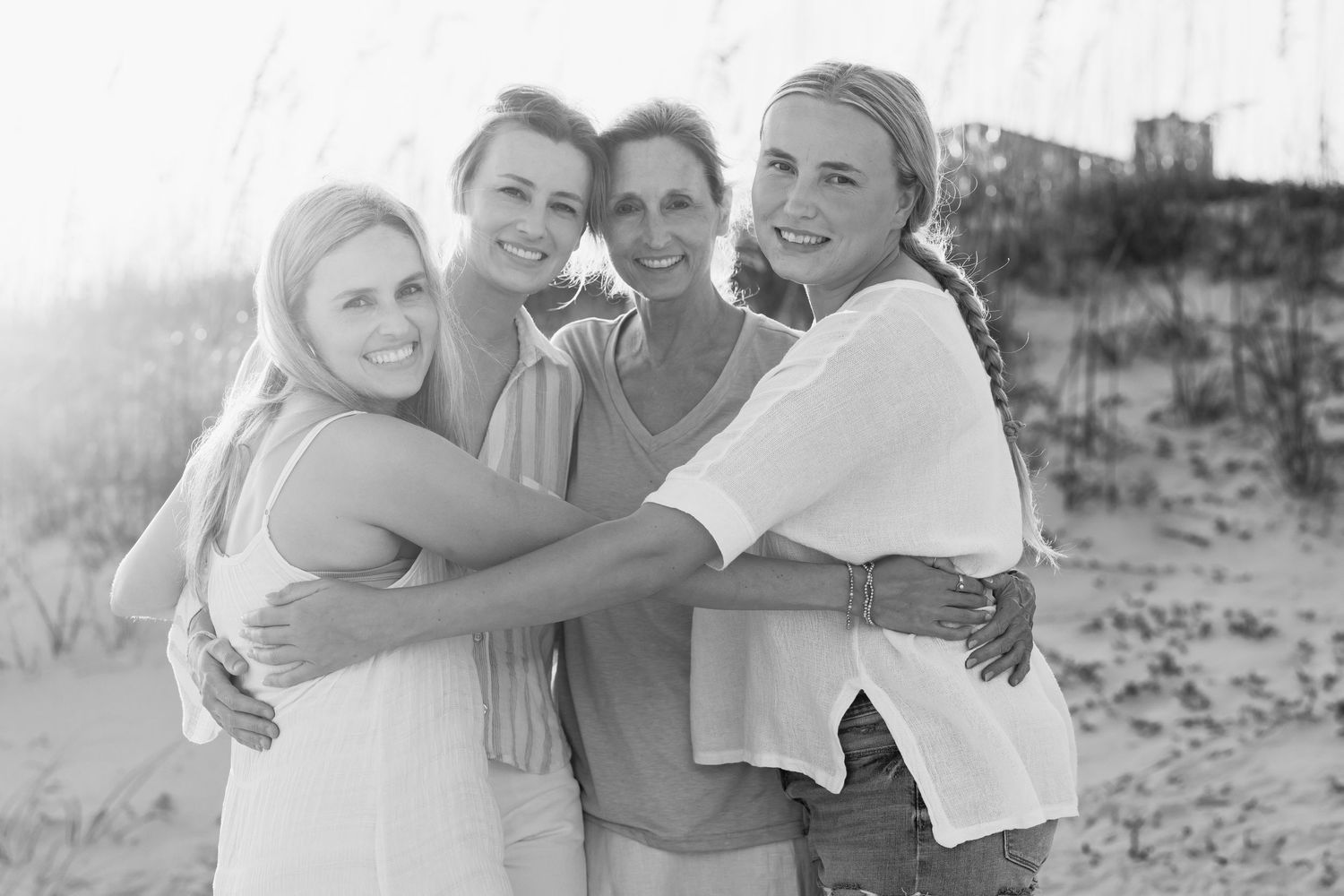 Four women hugging and smiling together on a beach in a black and white photo.