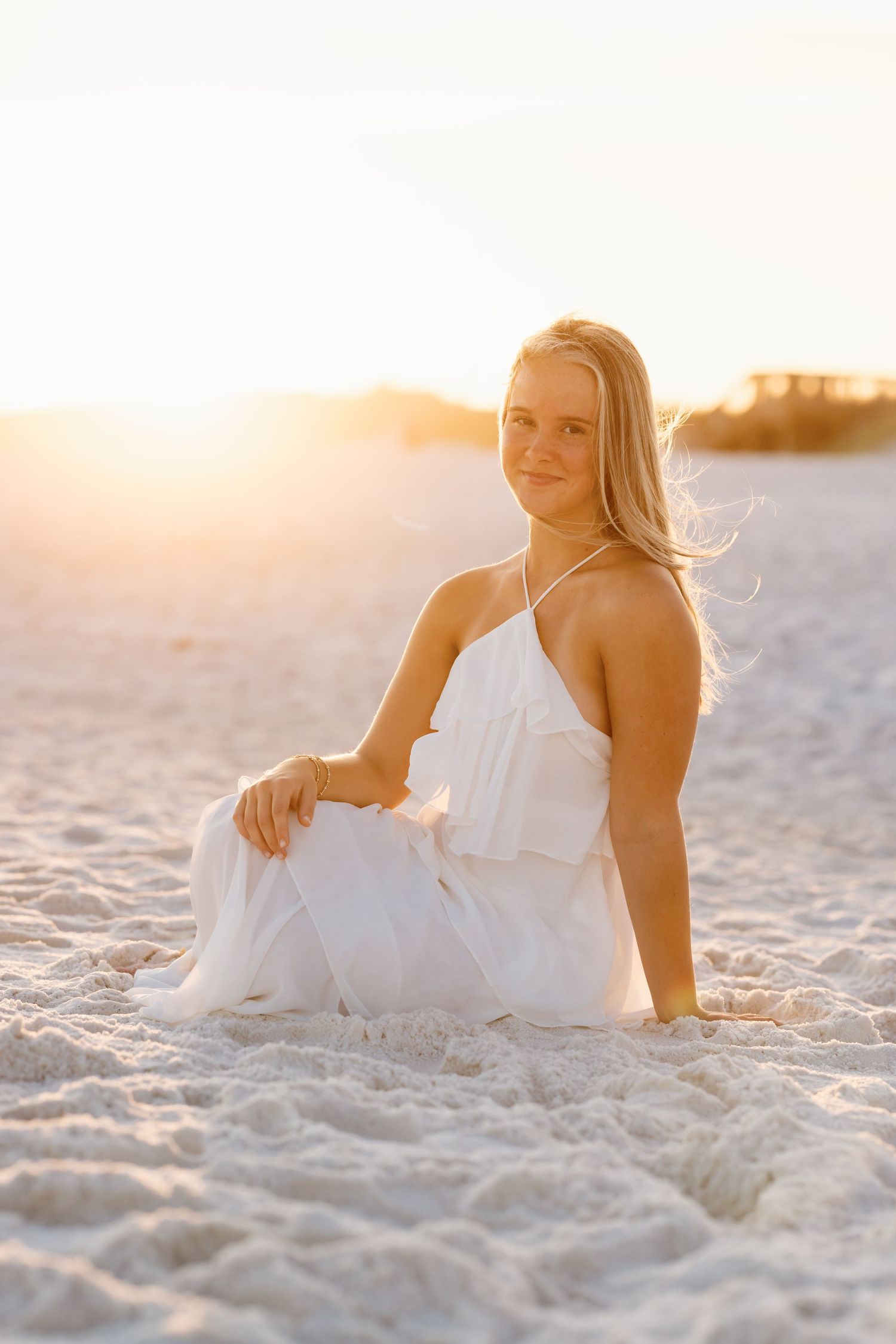 Blonde woman in white halter maxi dress sitting gracefully on sandy beach during golden hour sunset.