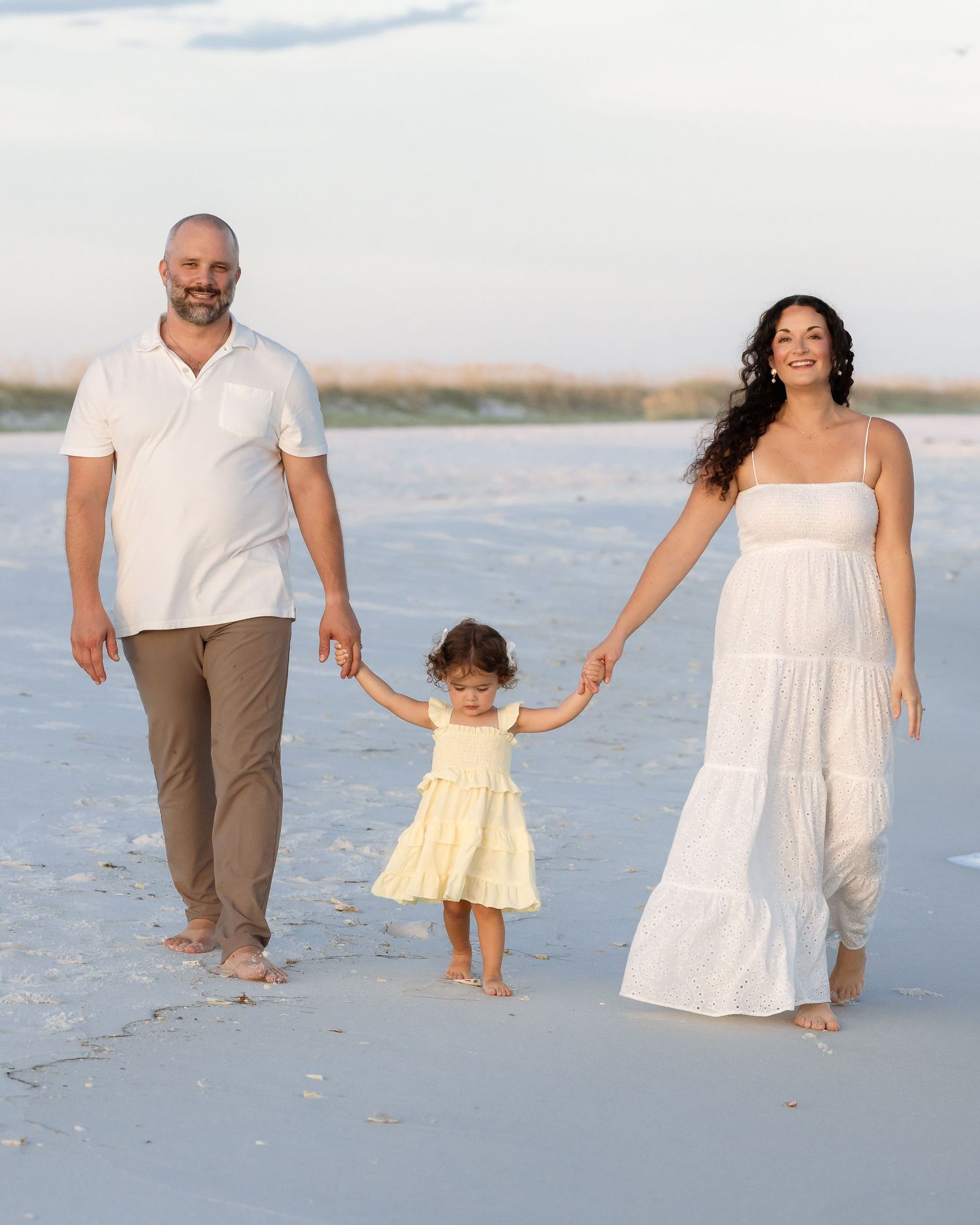 Family of three walking on beach, parents holding toddler girl's hands, wearing white and tan outfits.