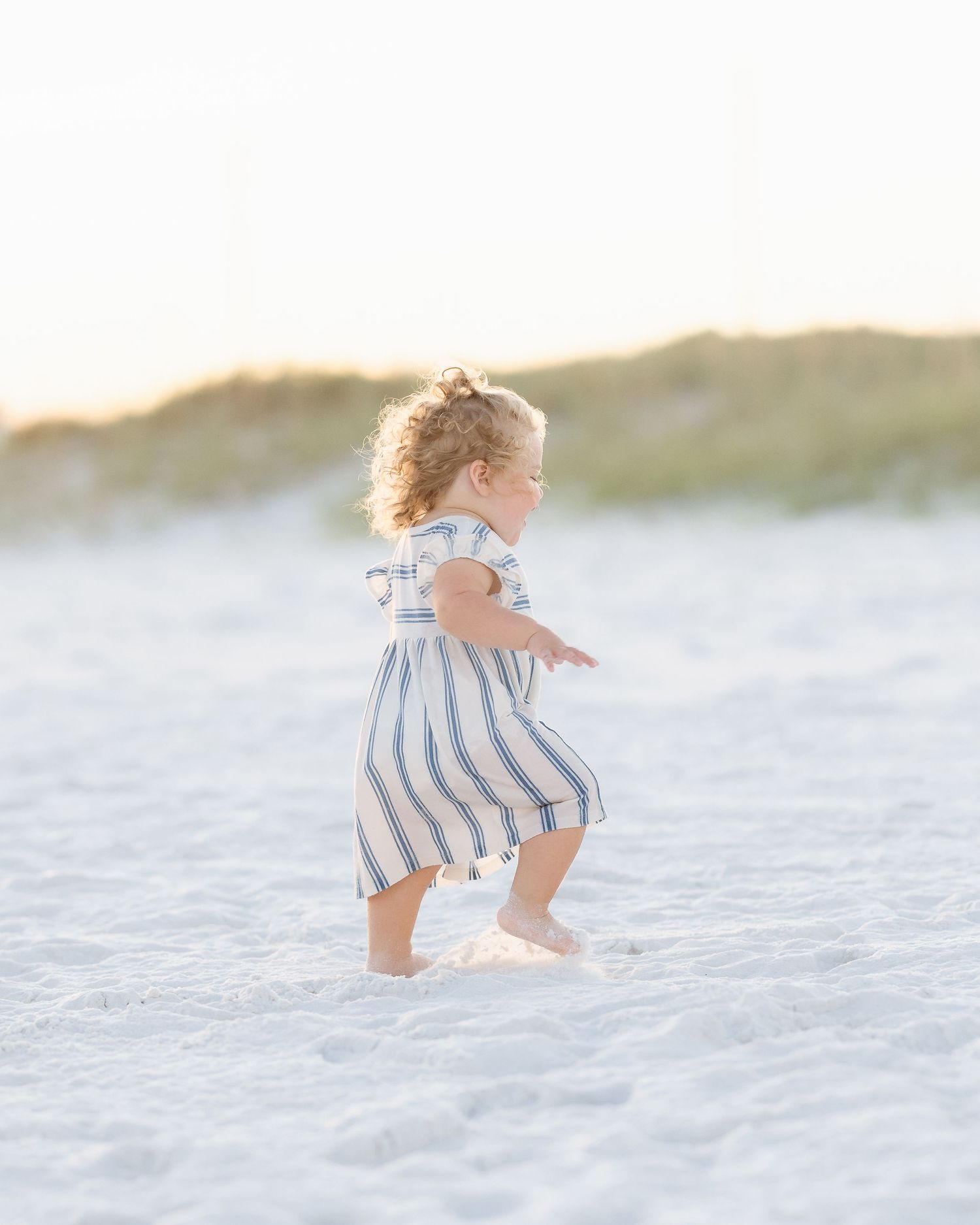 A toddler girl in a striped dress walks barefoot on a white sandy beach during golden hour.