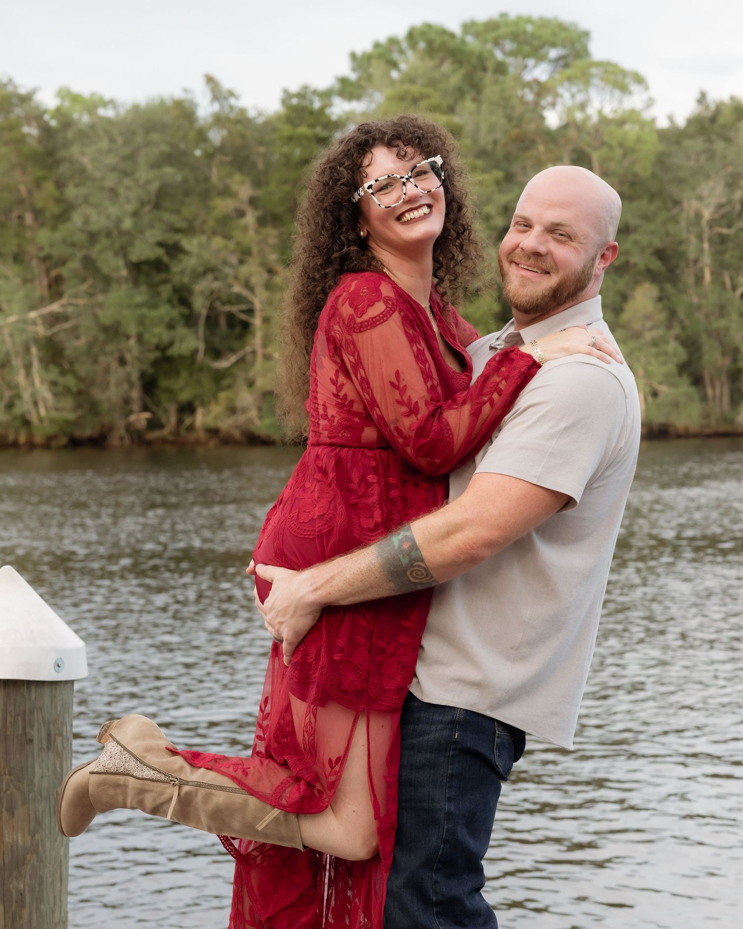 A smiling couple embraces on a waterfront dock, woman in red lace dress and boots, man in gray shirt and jeans.