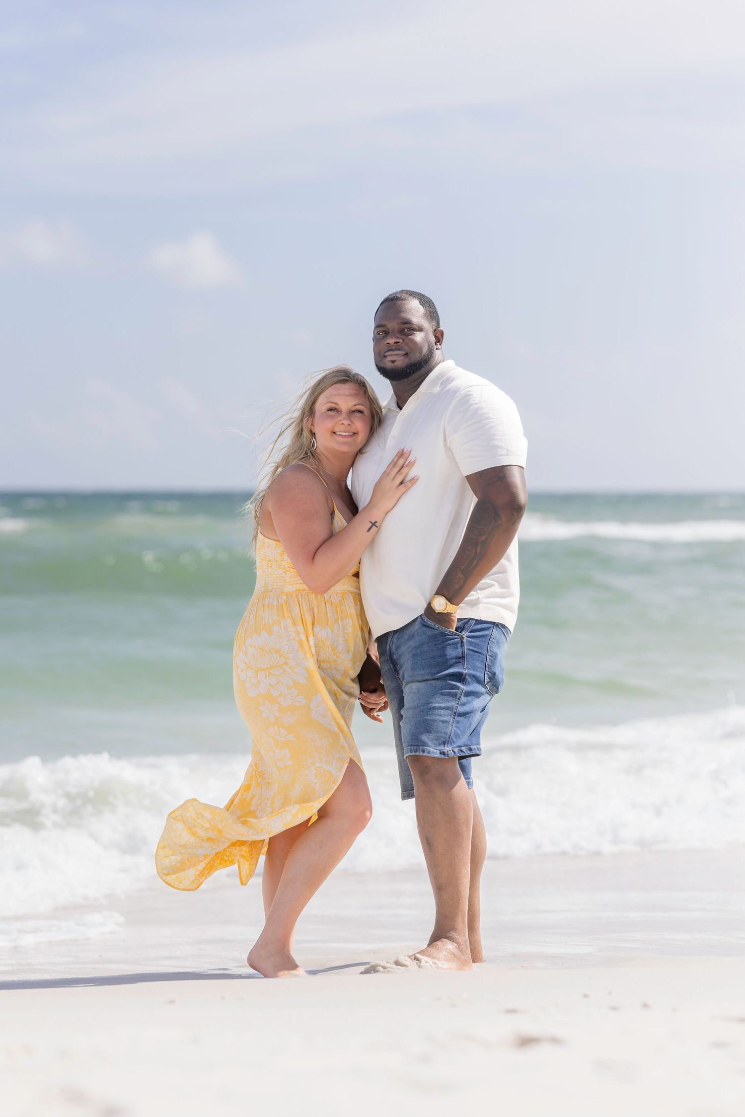Smiling interracial couple embracing barefoot on a white sandy beach with ocean waves behind them.
