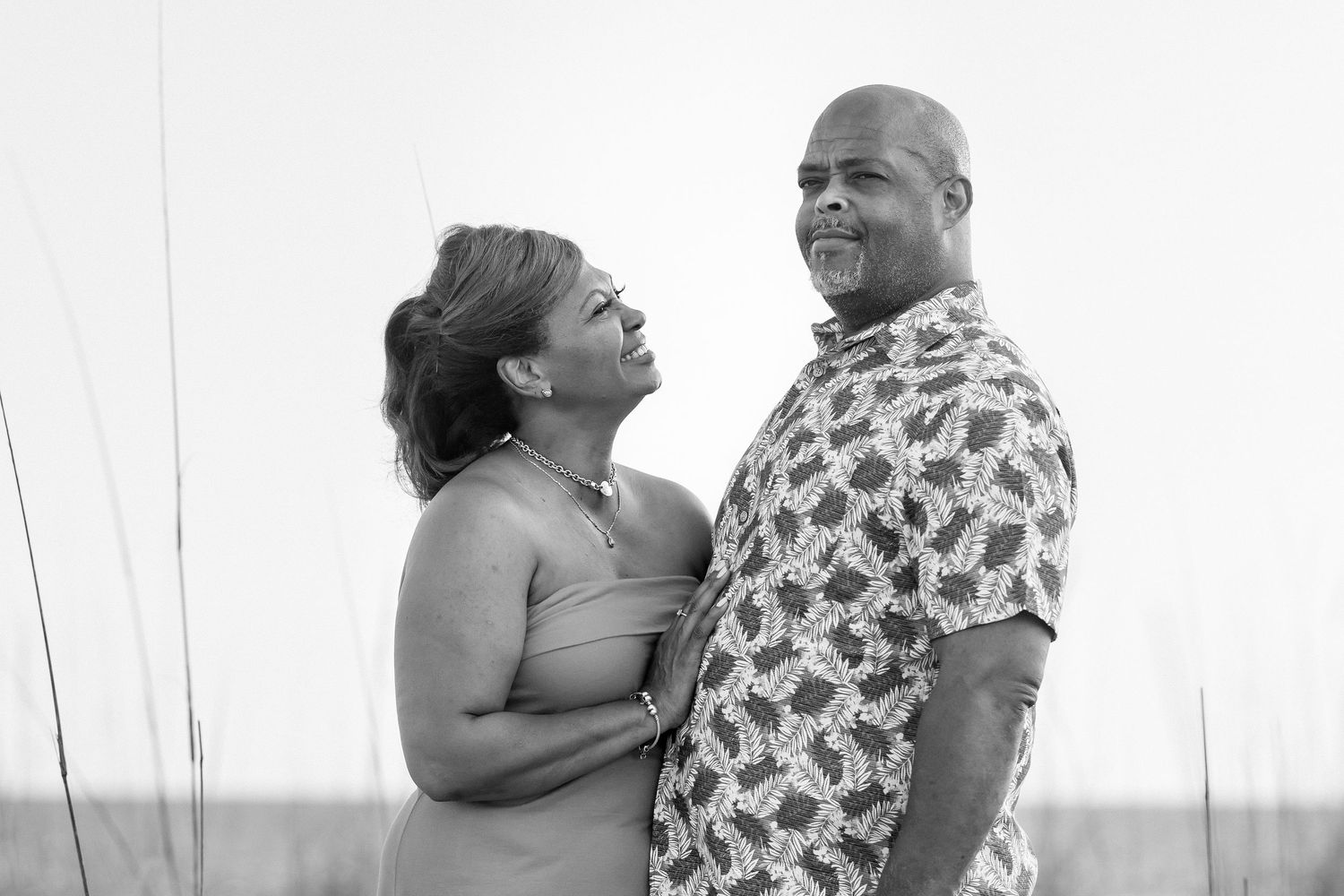 Black and white photo of a smiling couple embracing outdoors near tall grass, sharing a joyful moment together.