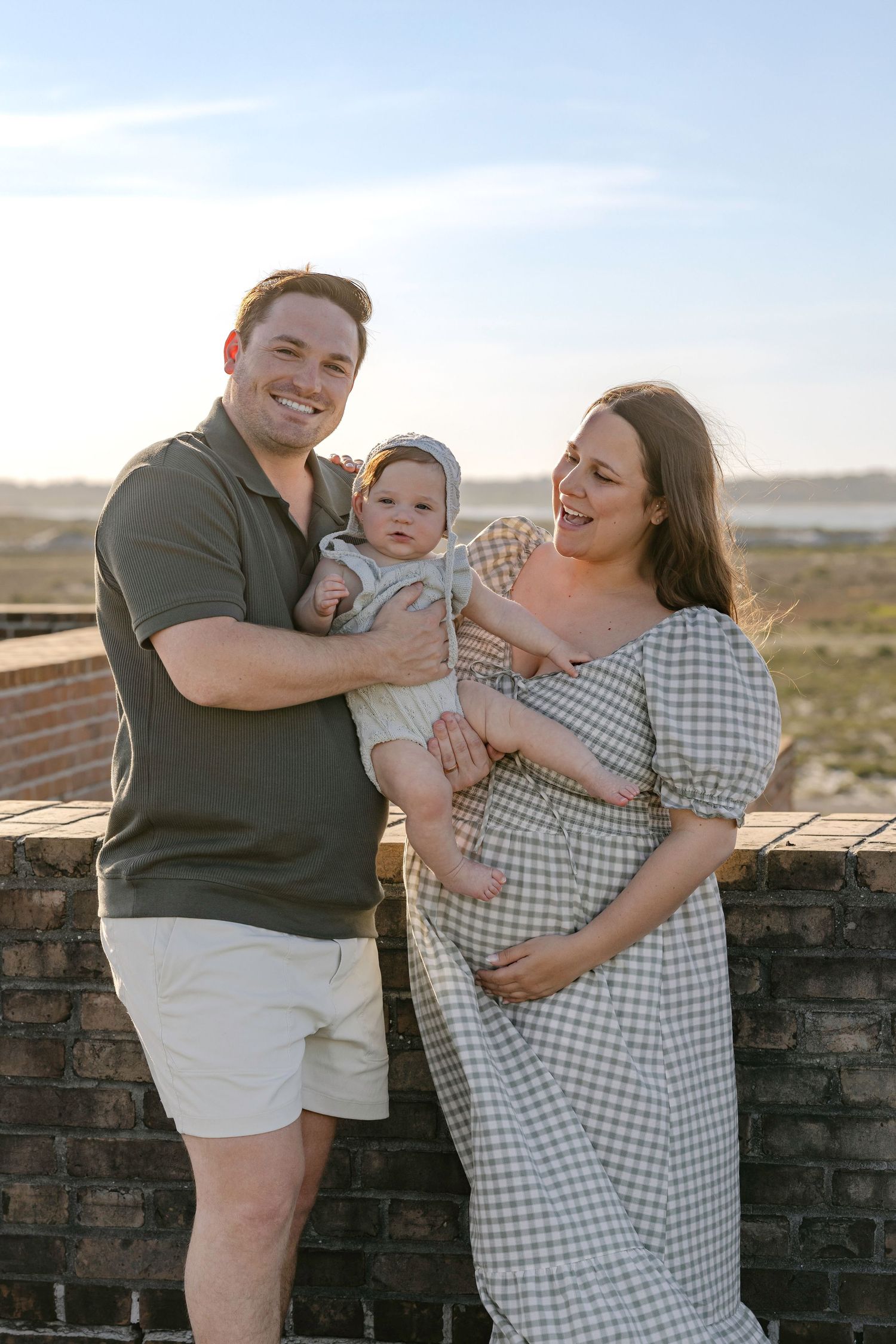 Happy expecting couple holding baby in bonnет against brick wall at golden hour outdoors.