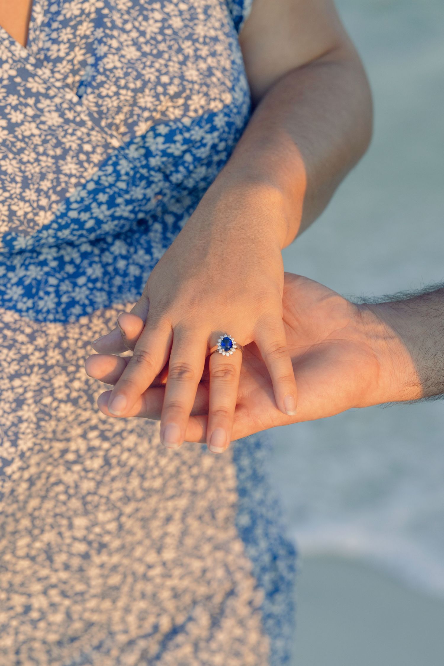 Close-up of couple holding hands showing sapphire engagement ring on woman's finger at beach.