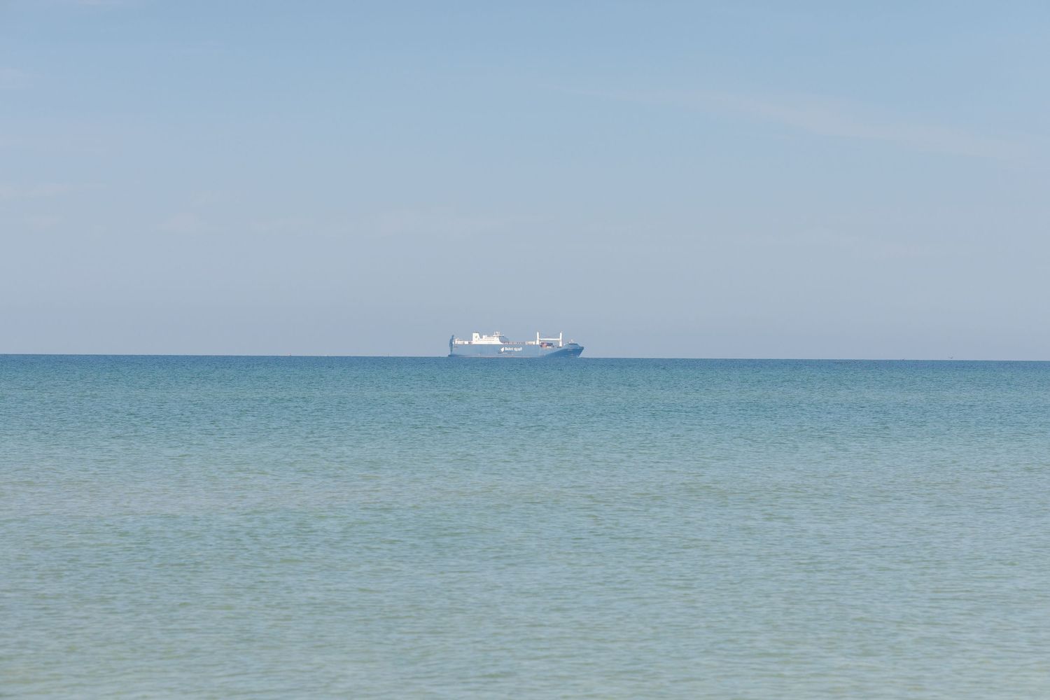 A serene turquoise ocean horizon with a cargo ship visible in the distance on a bright sunny day.