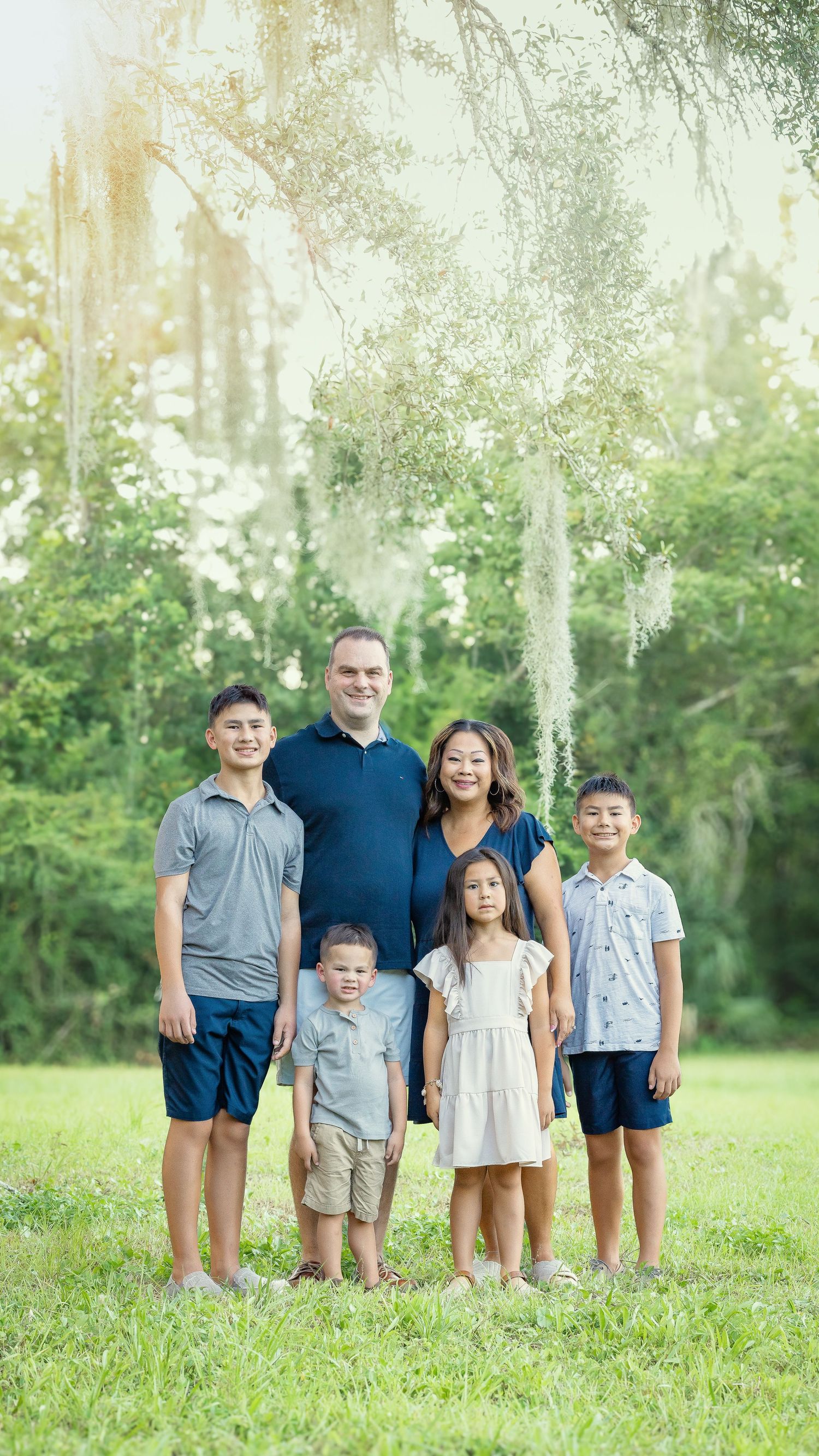 A family stands together outdoors on a grassy field with sunlit trees in the background for a pleasant portrait.