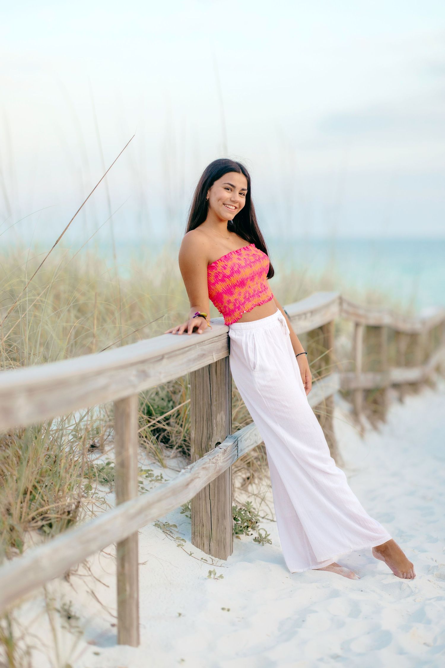 A figure in a coral pink top and white flowing pants poses on a wooden boardwalk by a beach with coastal dunes.
