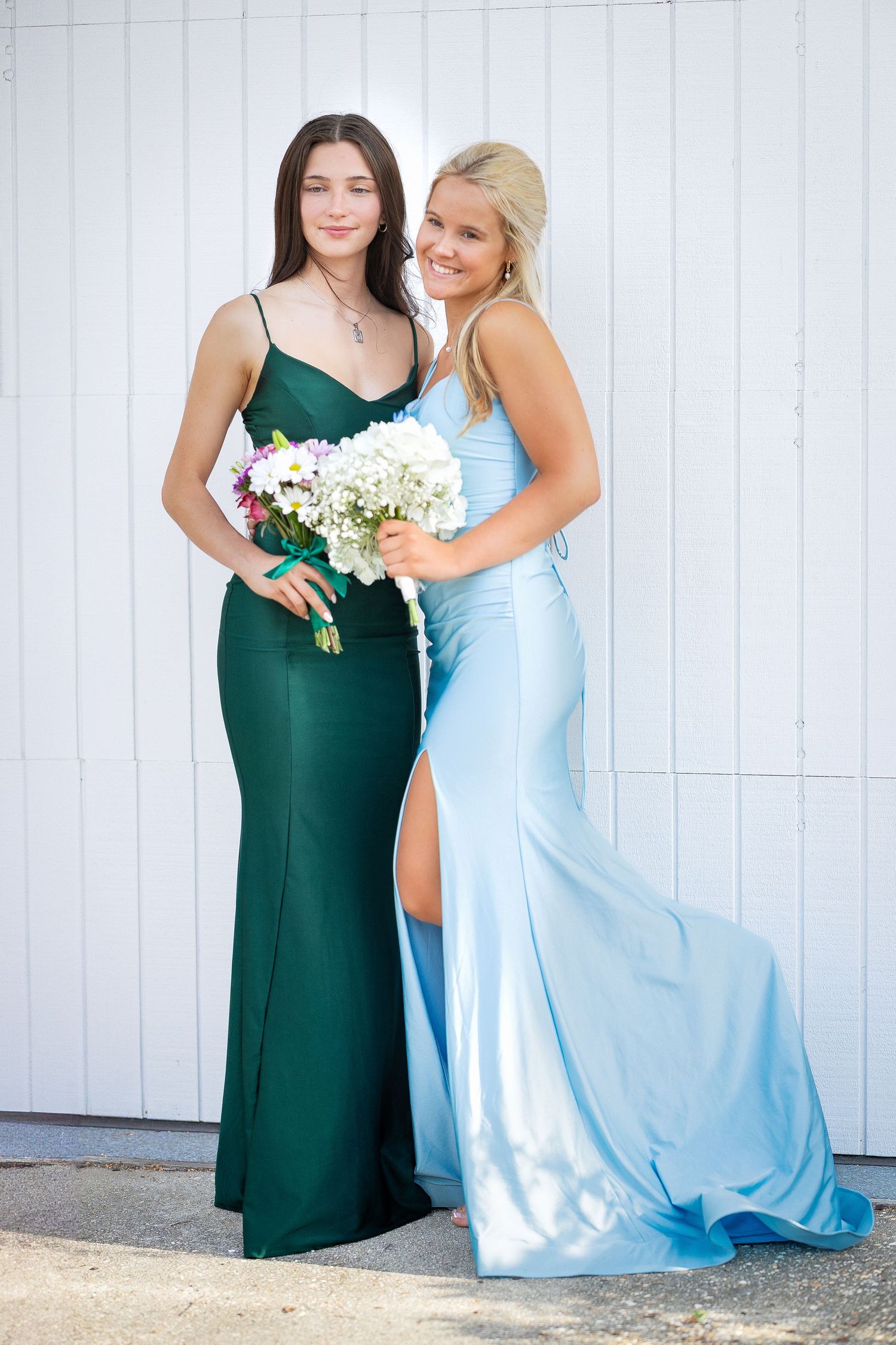 Two women pose together in elegant evening gowns - one in emerald green holding white flowers and one in light blue silk.