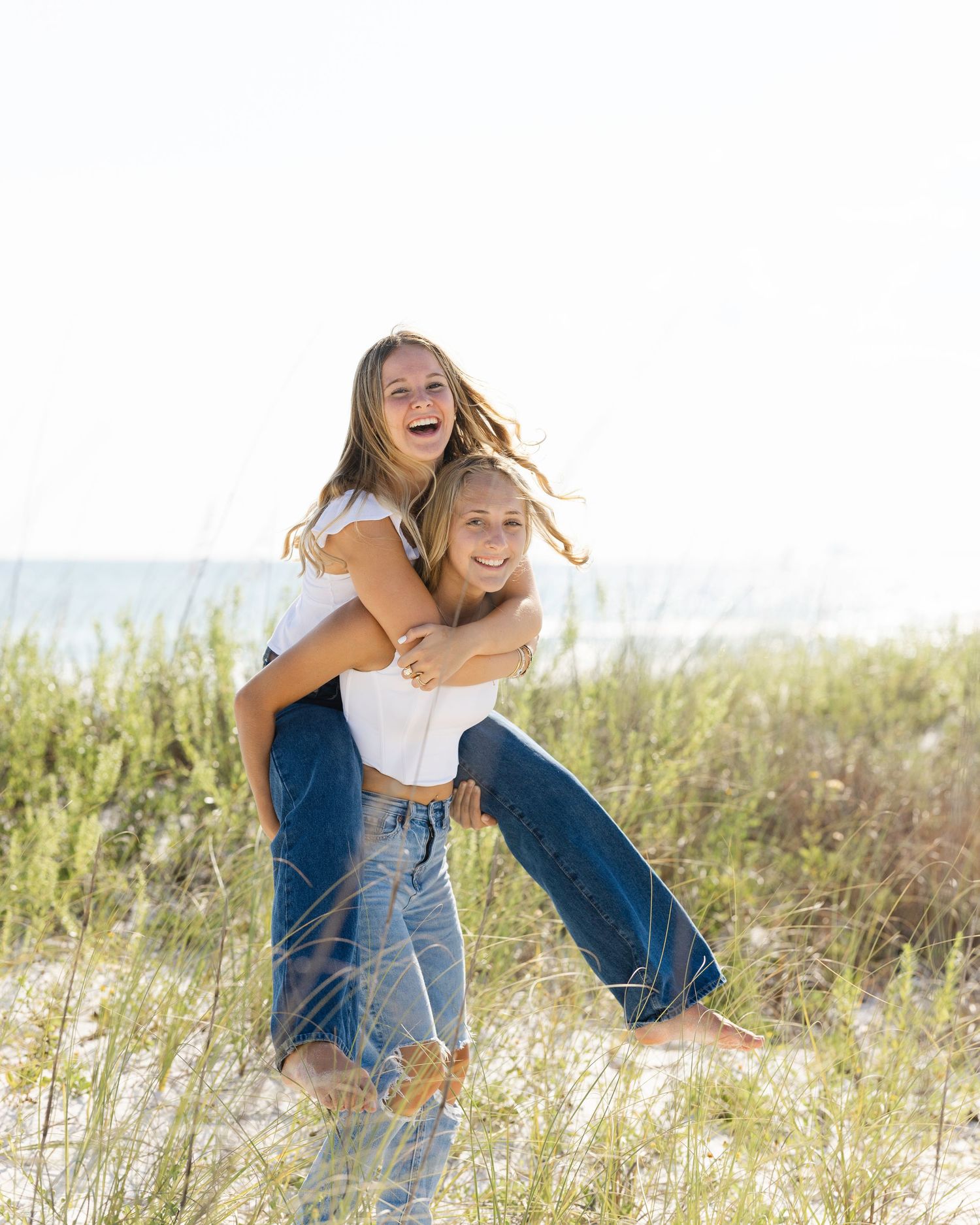 Two friends laugh together while giving a piggyback ride on sandy dunes near the ocean on a bright sunny day.