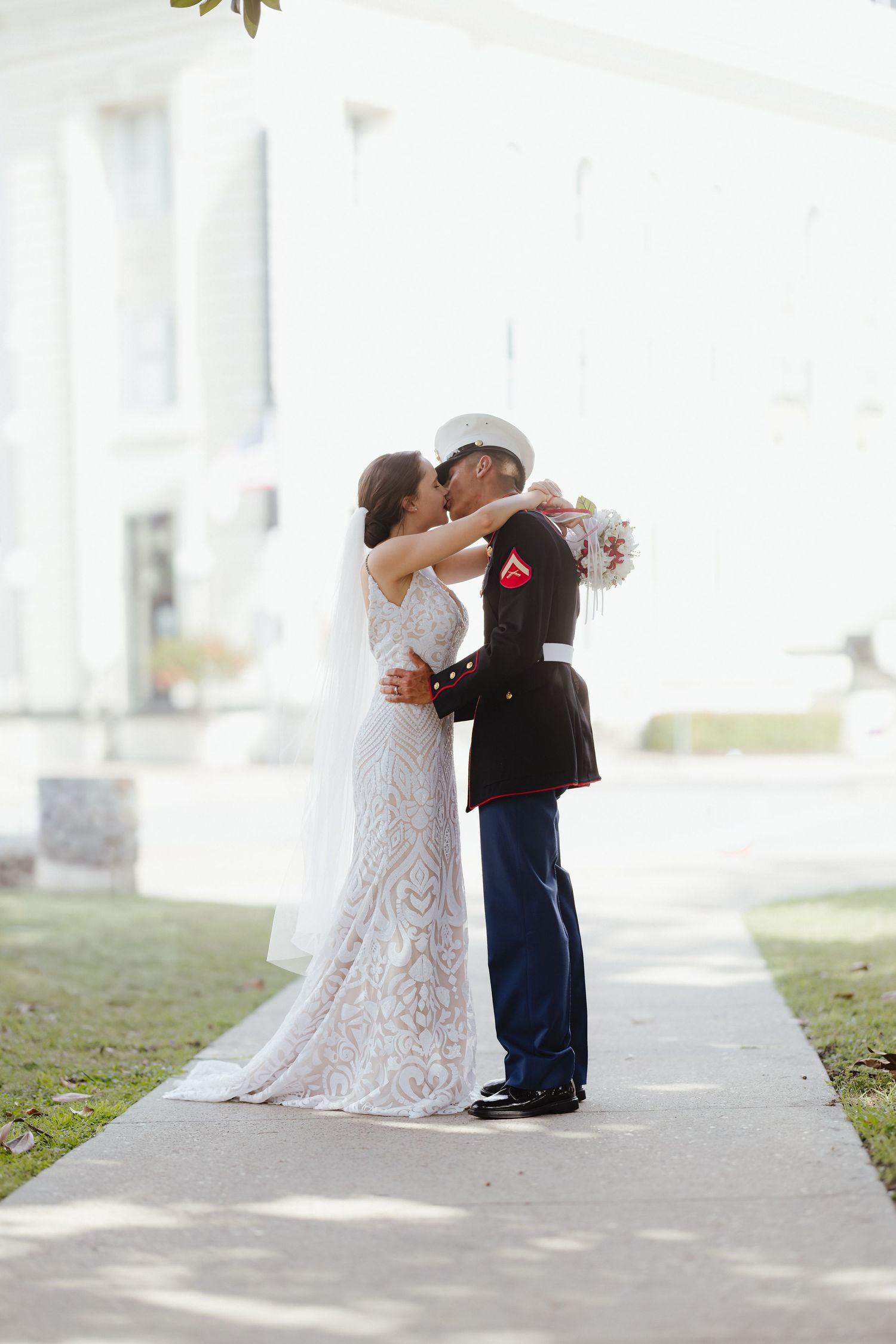 Marine groom in dress blues kissing bride in lace wedding gown on outdoor walkway.