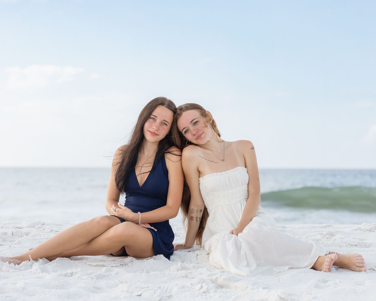 Two young women sitting together on sandy beach, wearing navy and white dresses, smiling at camera.