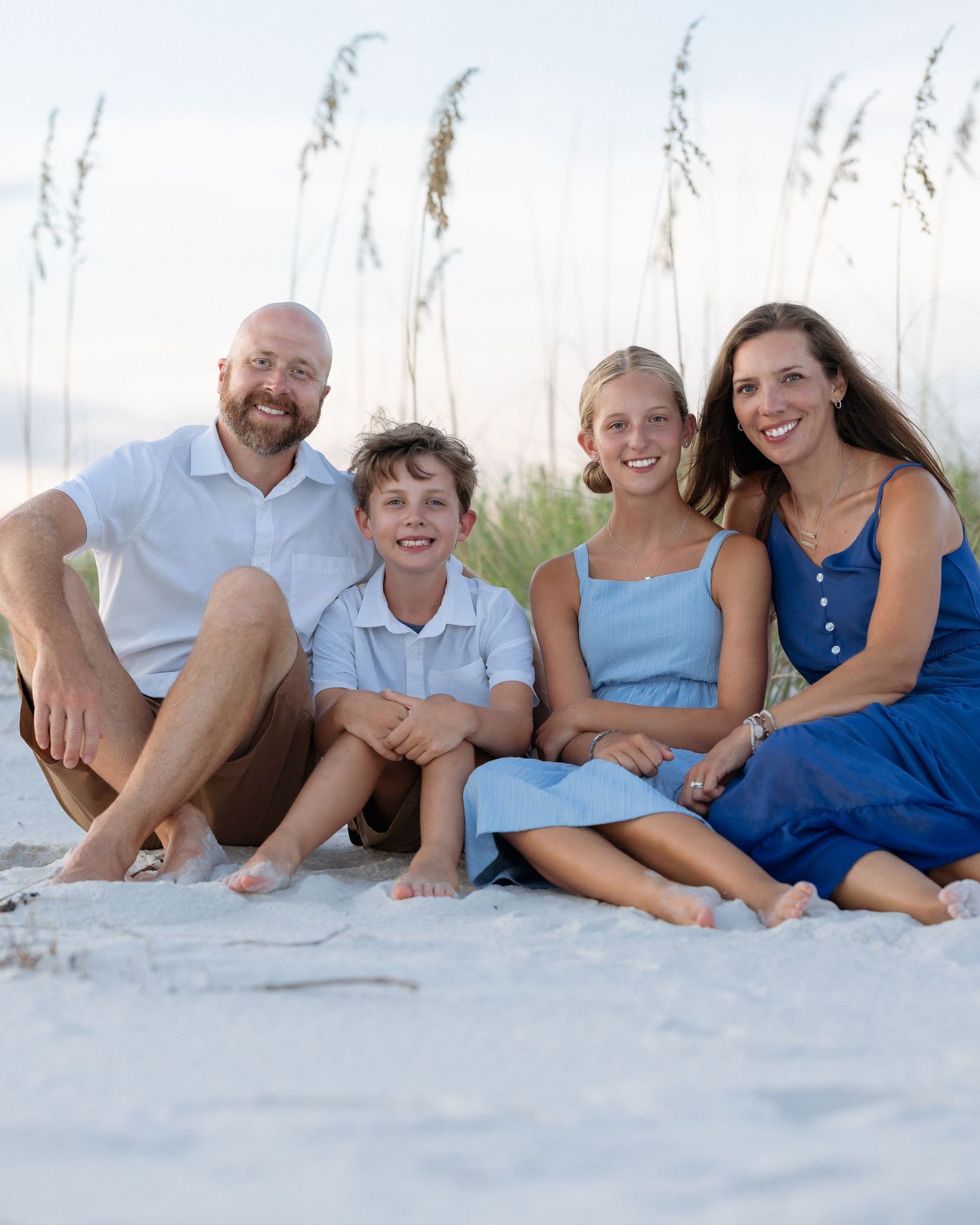 Happy family of four sitting together on a sandy beach with sea oats in background.