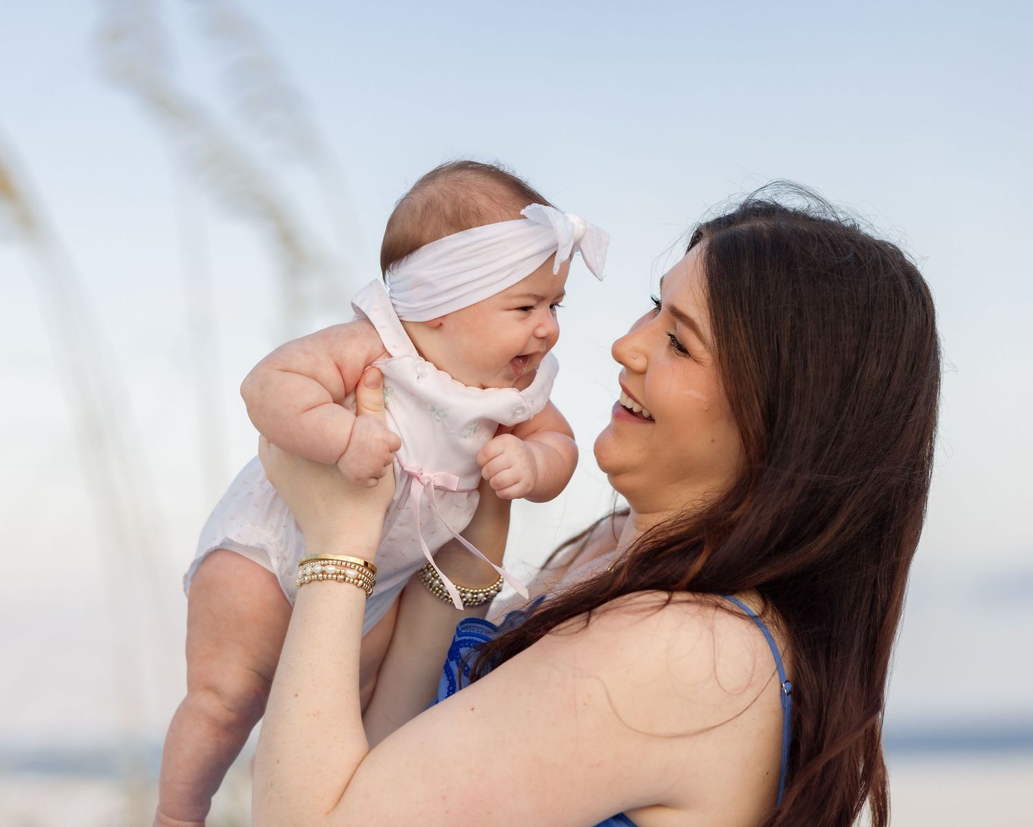 Young mother joyfully lifting her laughing baby girl wearing a white headband at the beach.