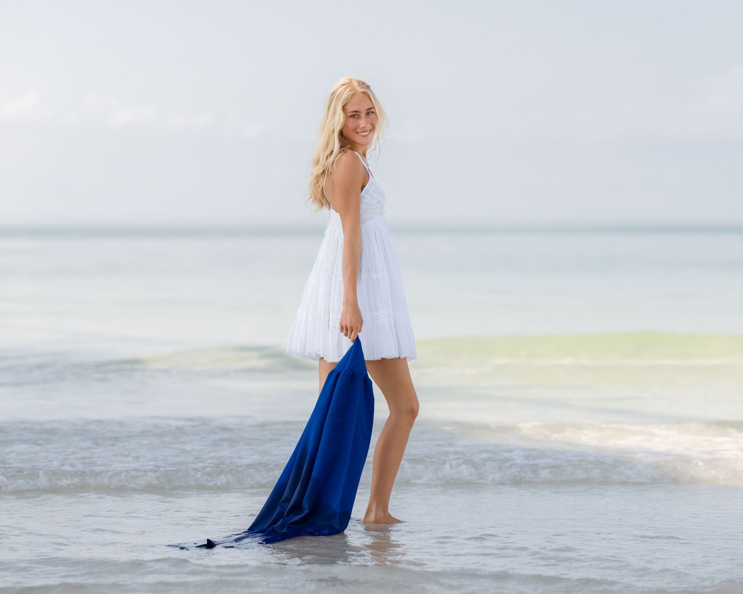 Blonde woman in white dress holding blue scarf wading in ocean waves on a sunny beach.