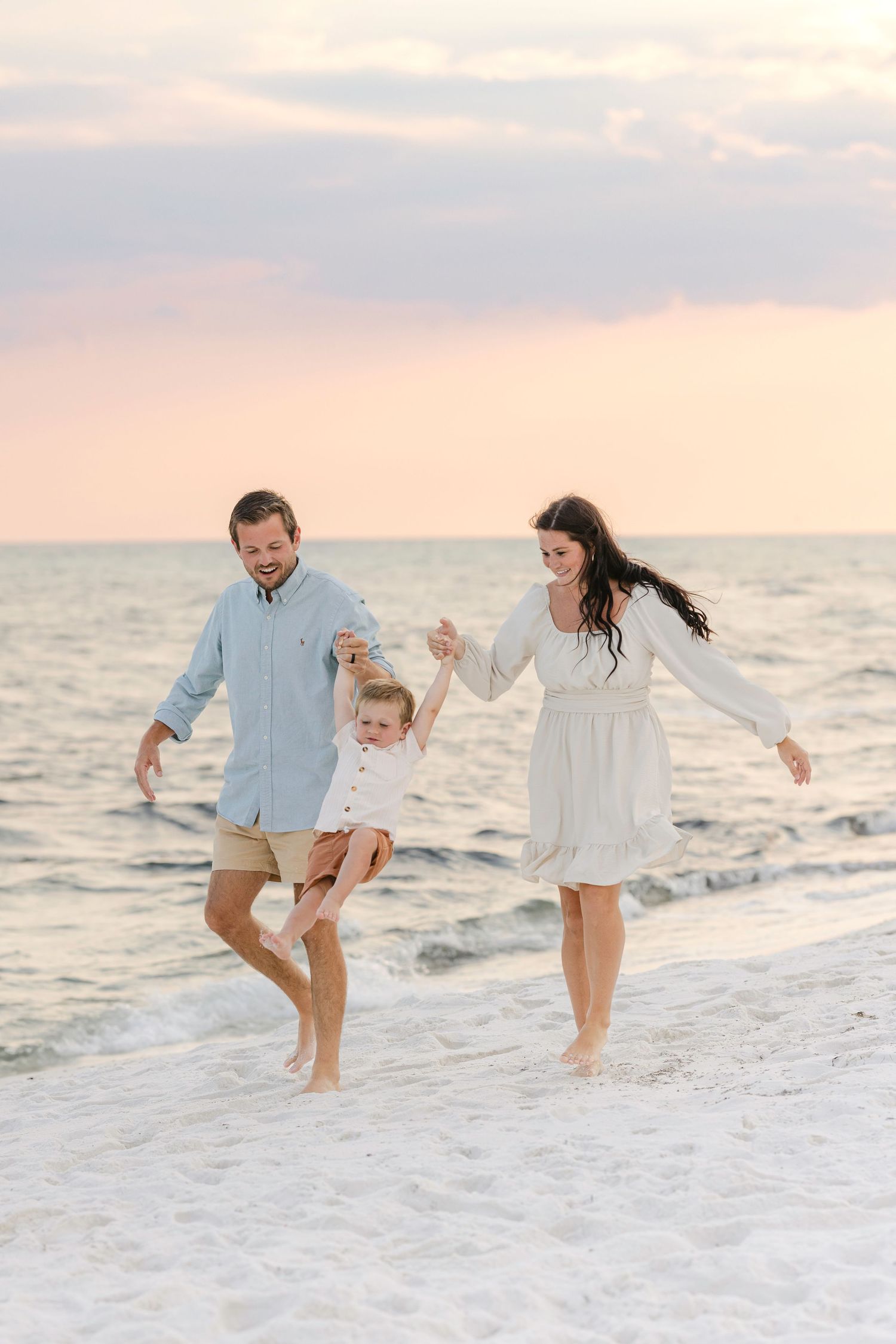 Family plays together on white sandy beach at sunset wearing light blue and white outfits.
