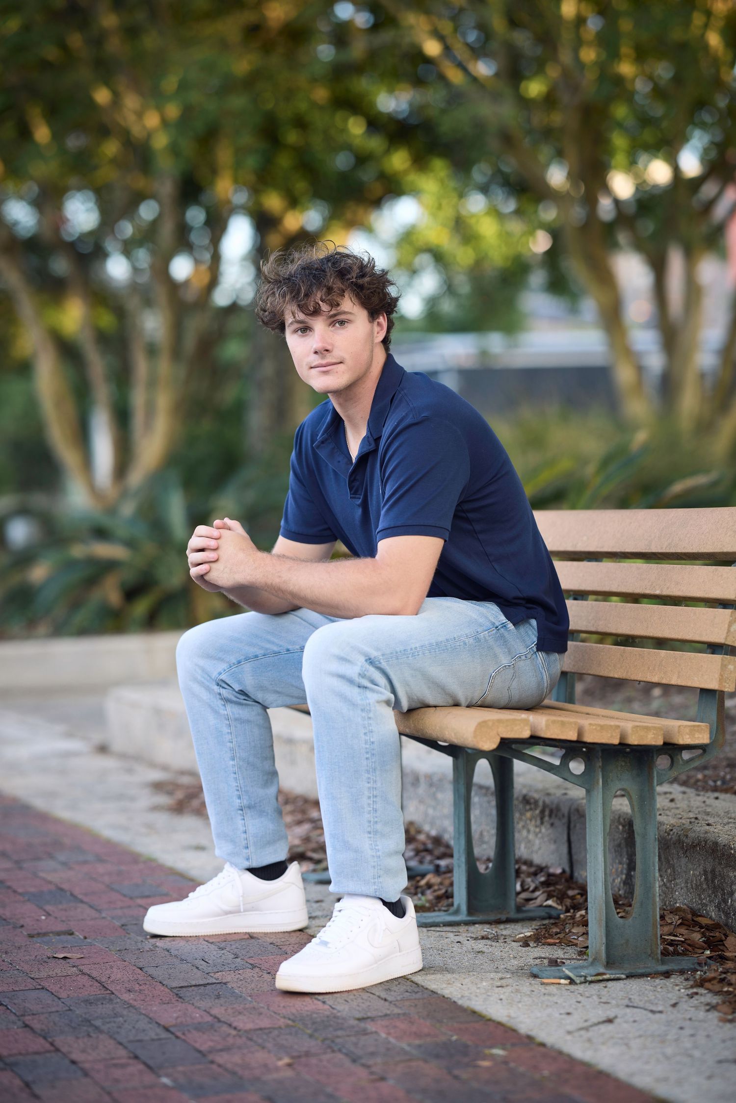Person in navy polo and jeans sitting on wooden bench in outdoor park setting.