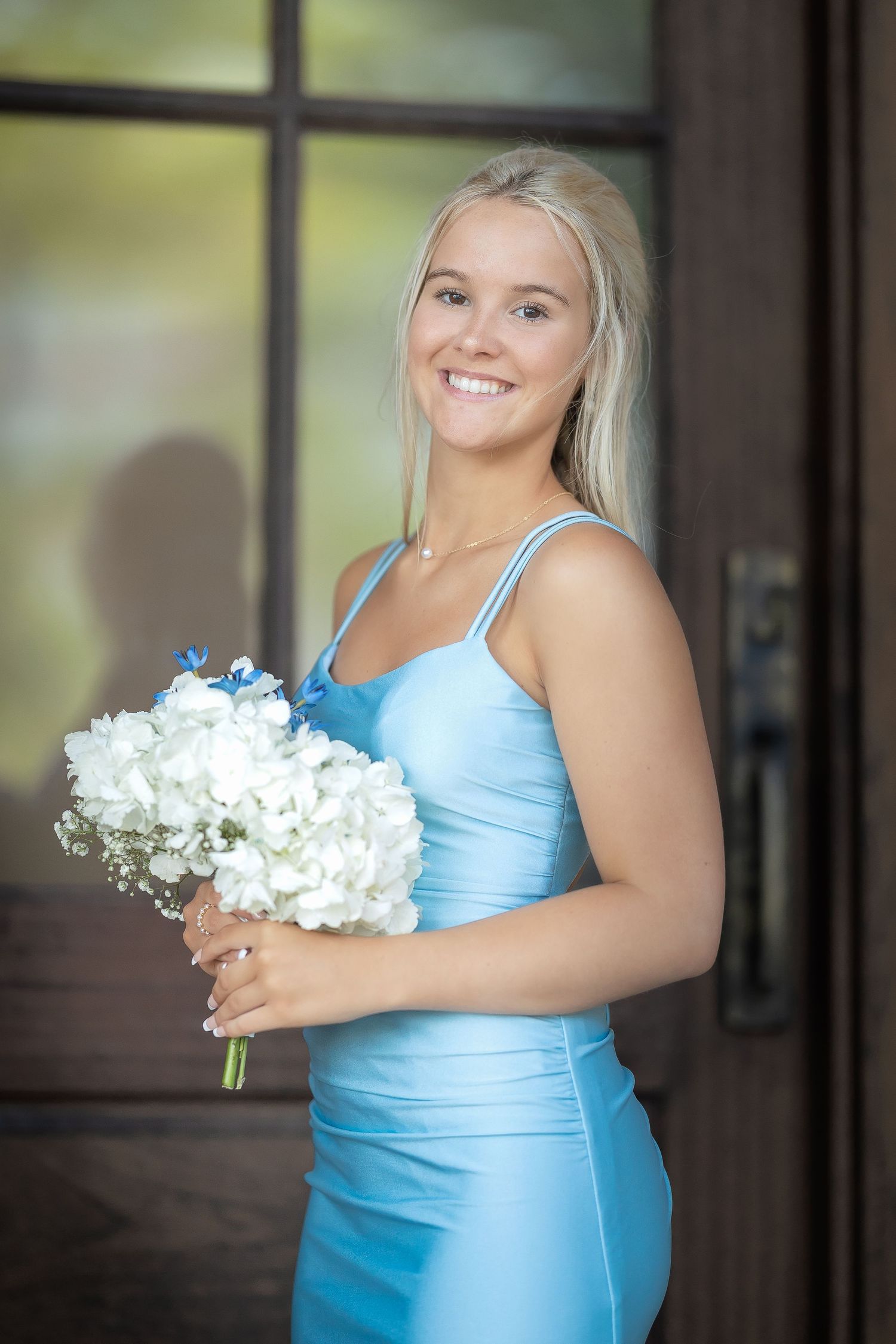 A person in a light blue bridesmaid dress holds white flowers while posing in front of a window.