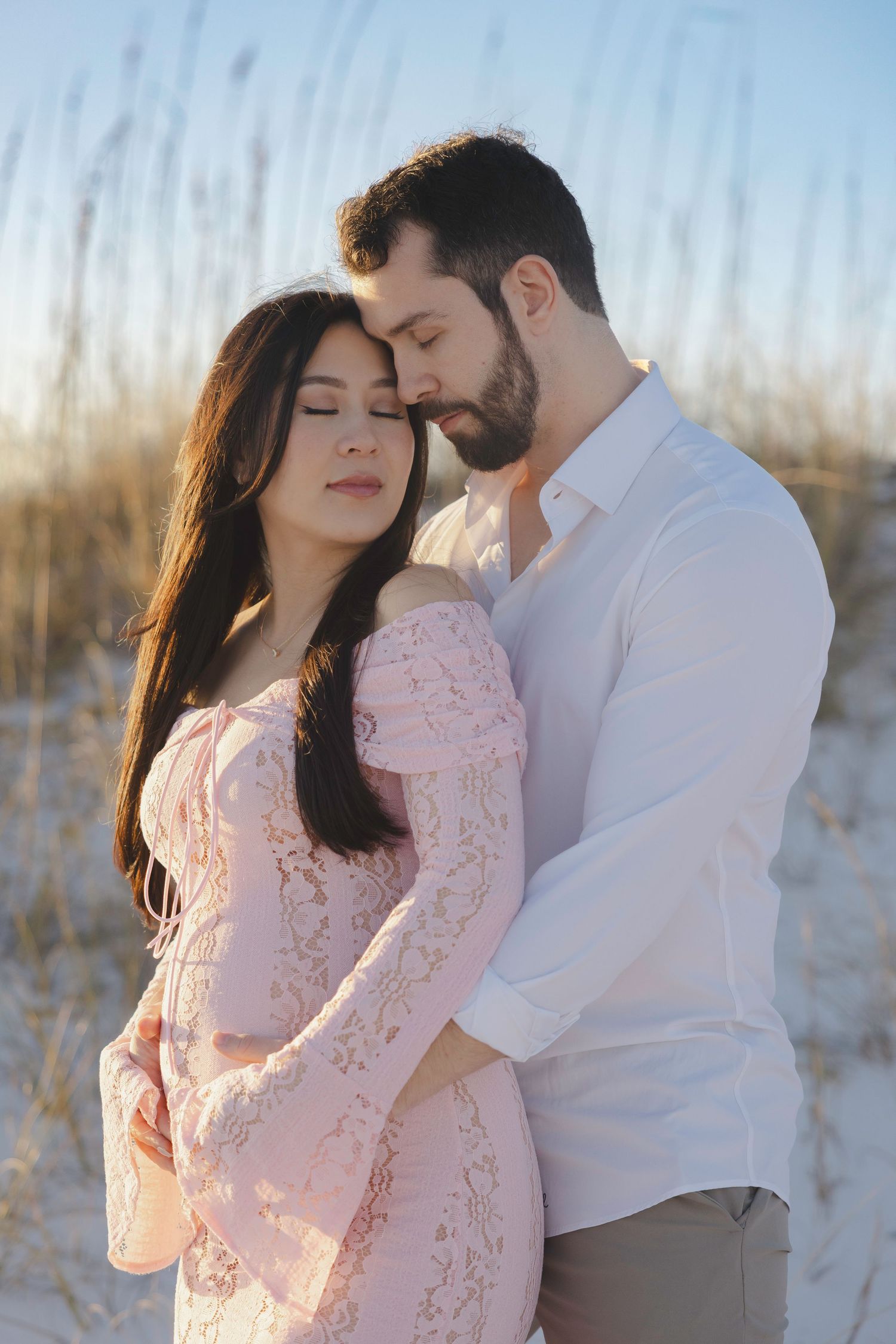 Romantic couple in pink lace and white embracing on sandy beach during golden hour sunset.