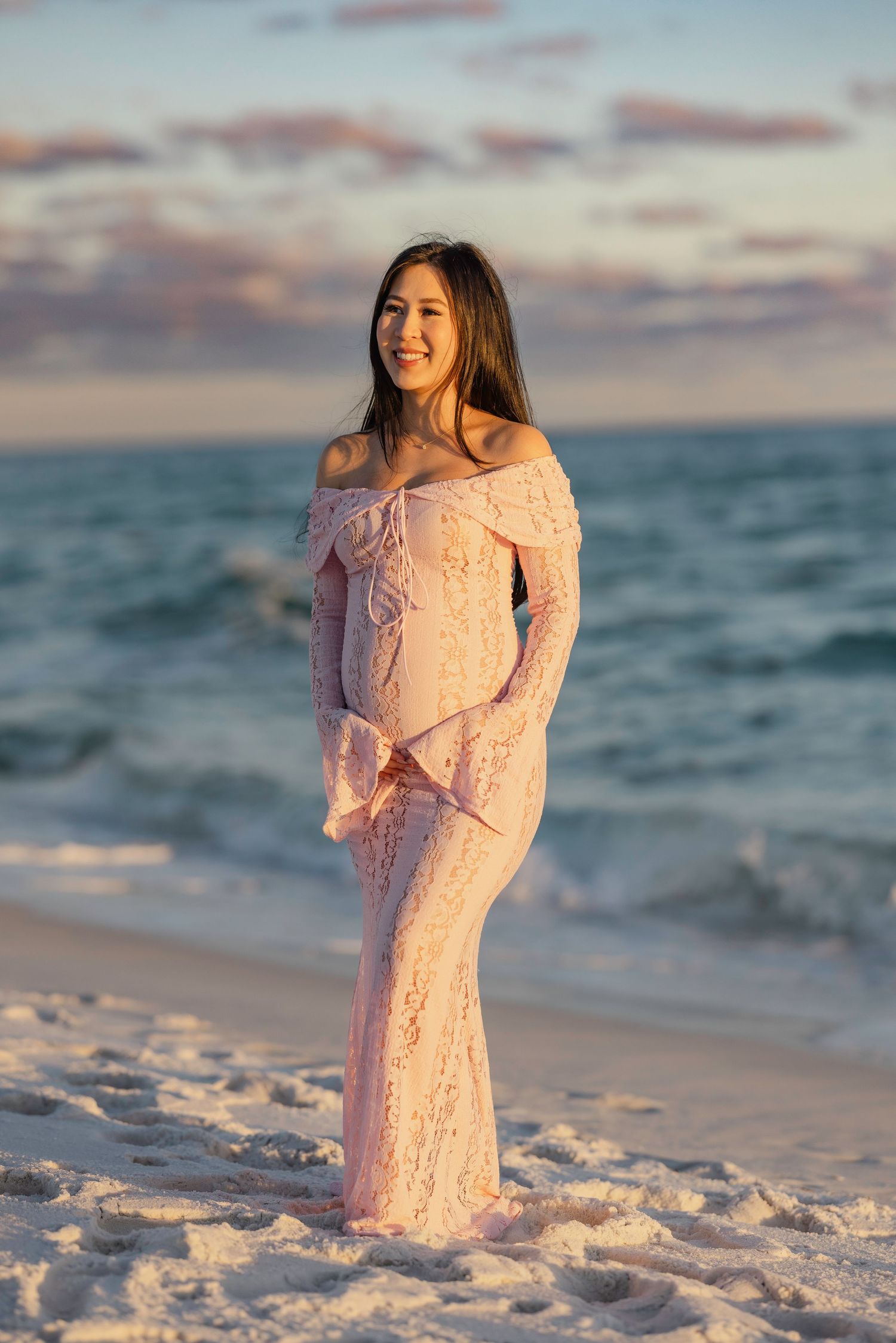 Someone wearing a pink lace dress poses gracefully on a sandy beach at sunset with ocean waves in the background.