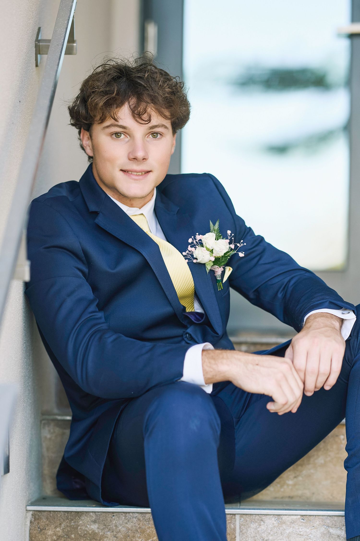 A person in a navy blue suit with yellow tie and boutonniere sits casually on stone steps at a formal event.