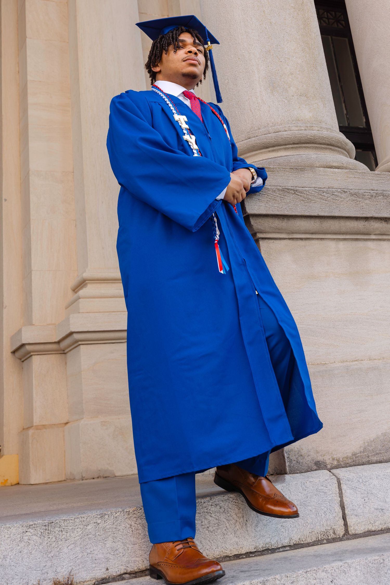Young man in blue graduation cap and gown standing on steps near stone columns, wearing brown dress shoes.