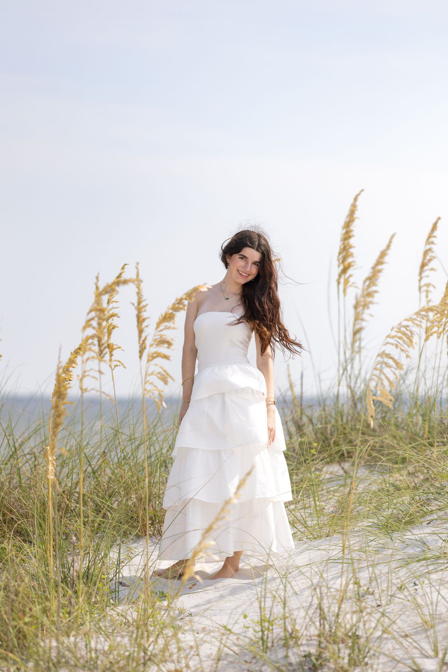 A woman in a white tiered dress stands barefoot on sandy beach dunes surrounded by golden sea oats grass.