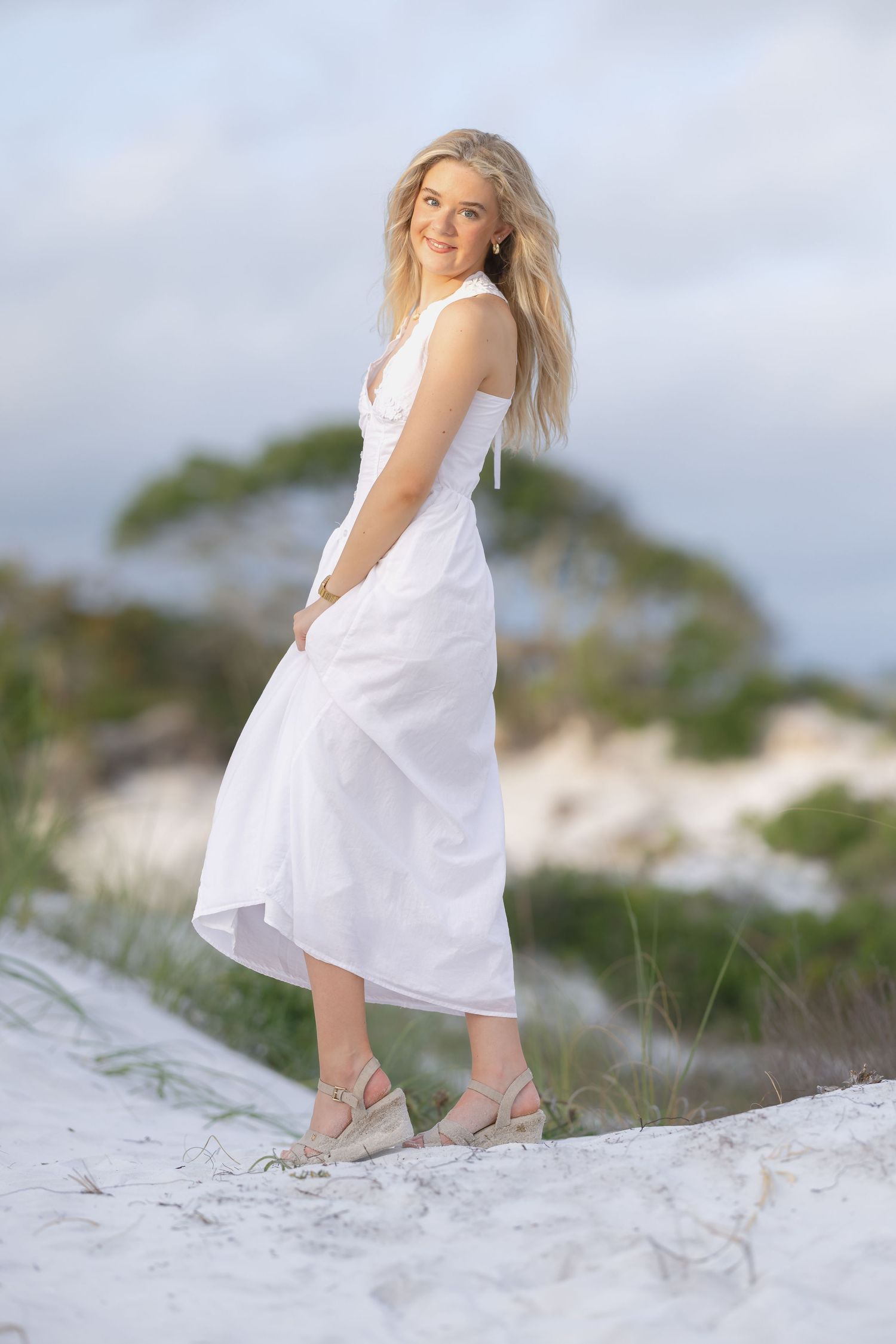 Smiling blonde woman in white midi dress and wedge sandals standing on white sand dunes with coastal vegetation.
