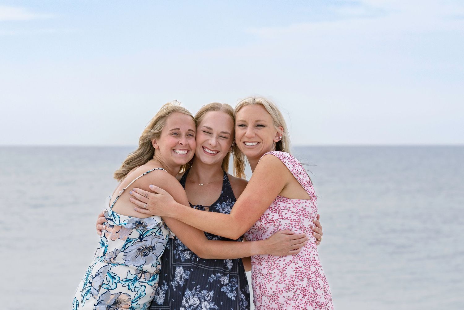 Three smiling blonde women hugging each other on a sunny beach with calm ocean water in the background.