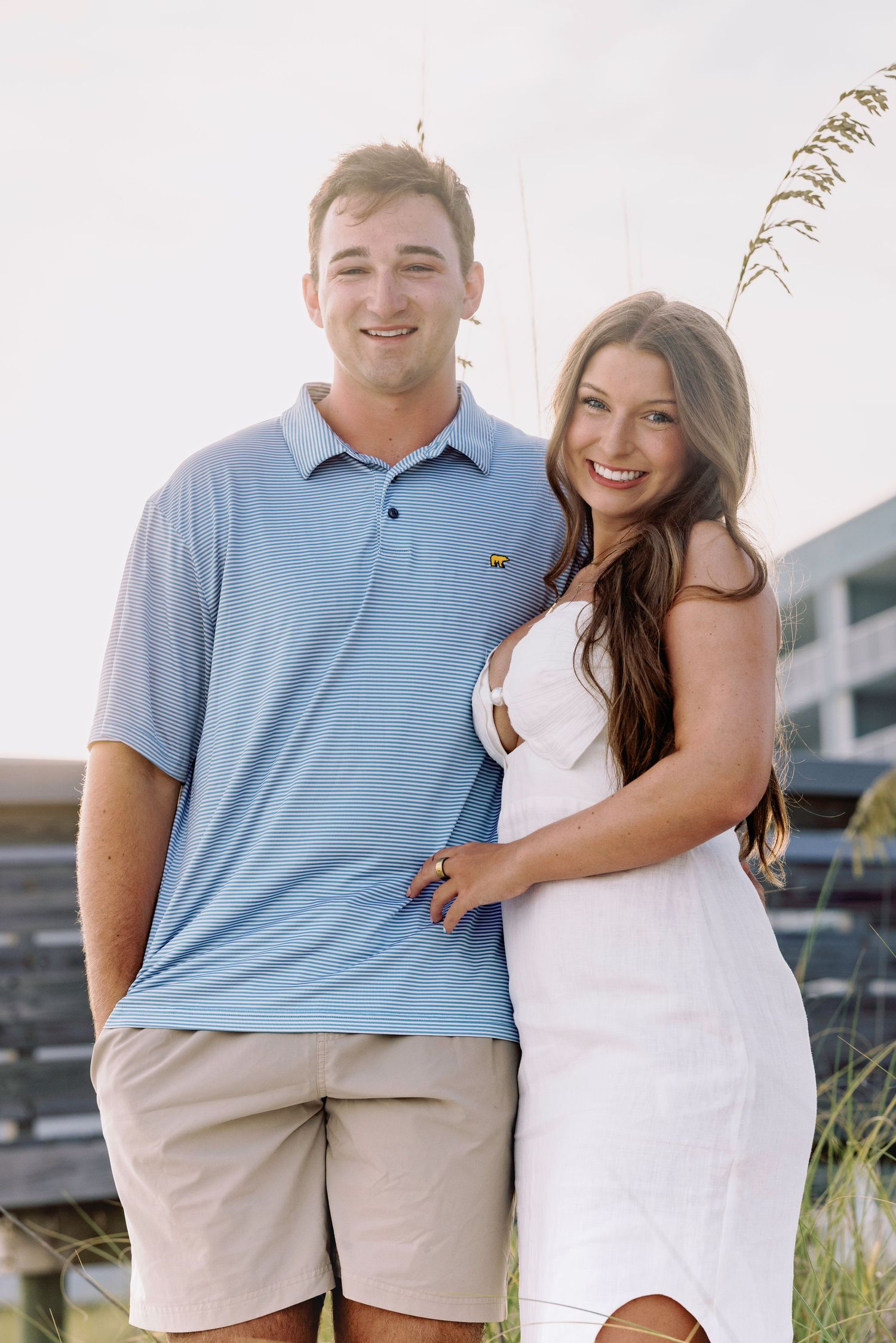 Smiling couple posing outdoors near beach dunes, man in blue polo and khaki shorts with woman in white dress.