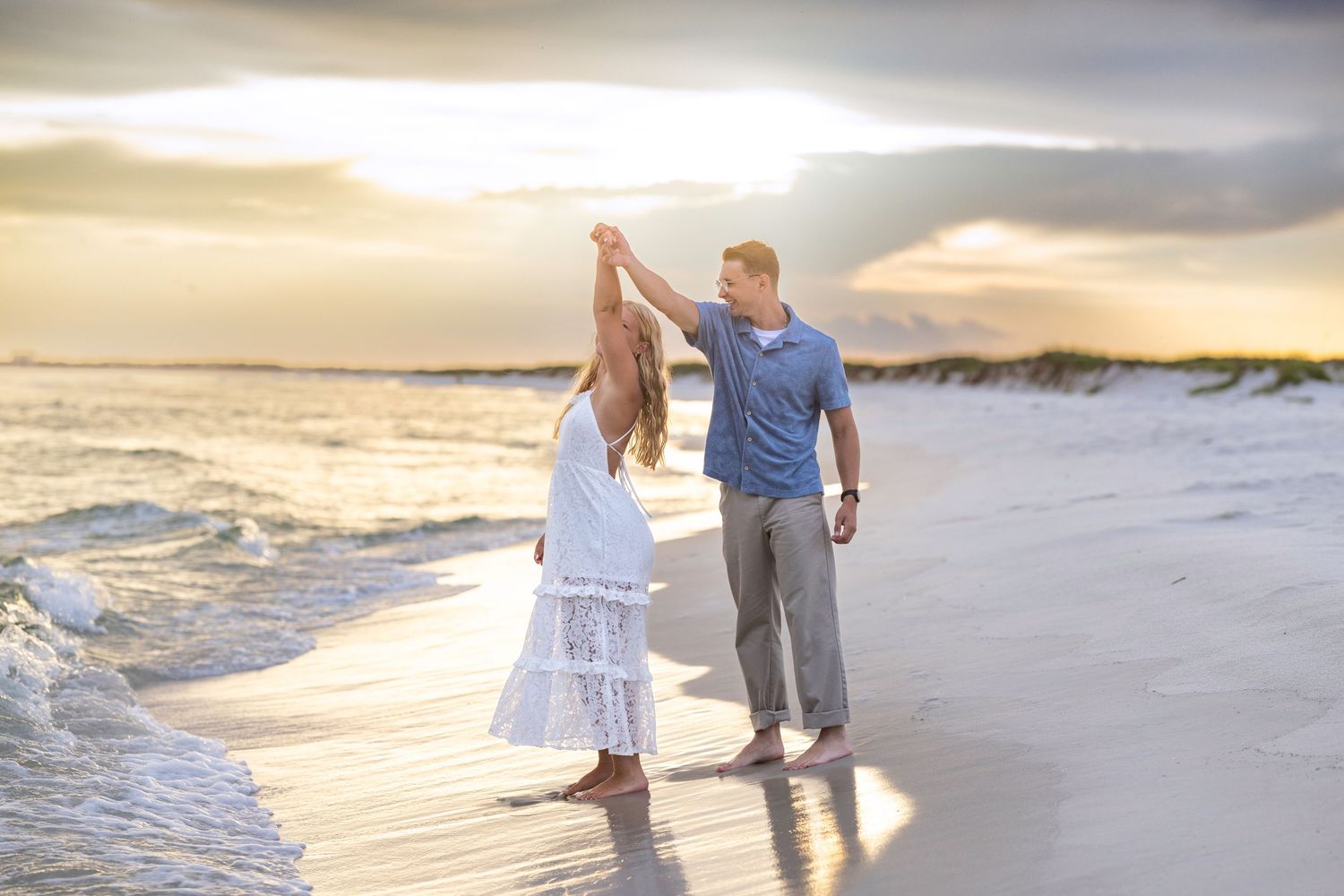 Couple dancing on a sandy beach at sunset, waves crashing nearby, woman in white dress, man in blue shirt.