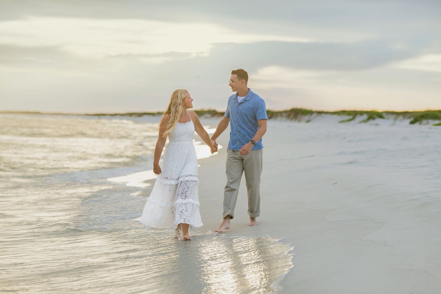 Couple holding hands walking barefoot on a sandy beach at sunset, woman in white dress, man in blue shirt.