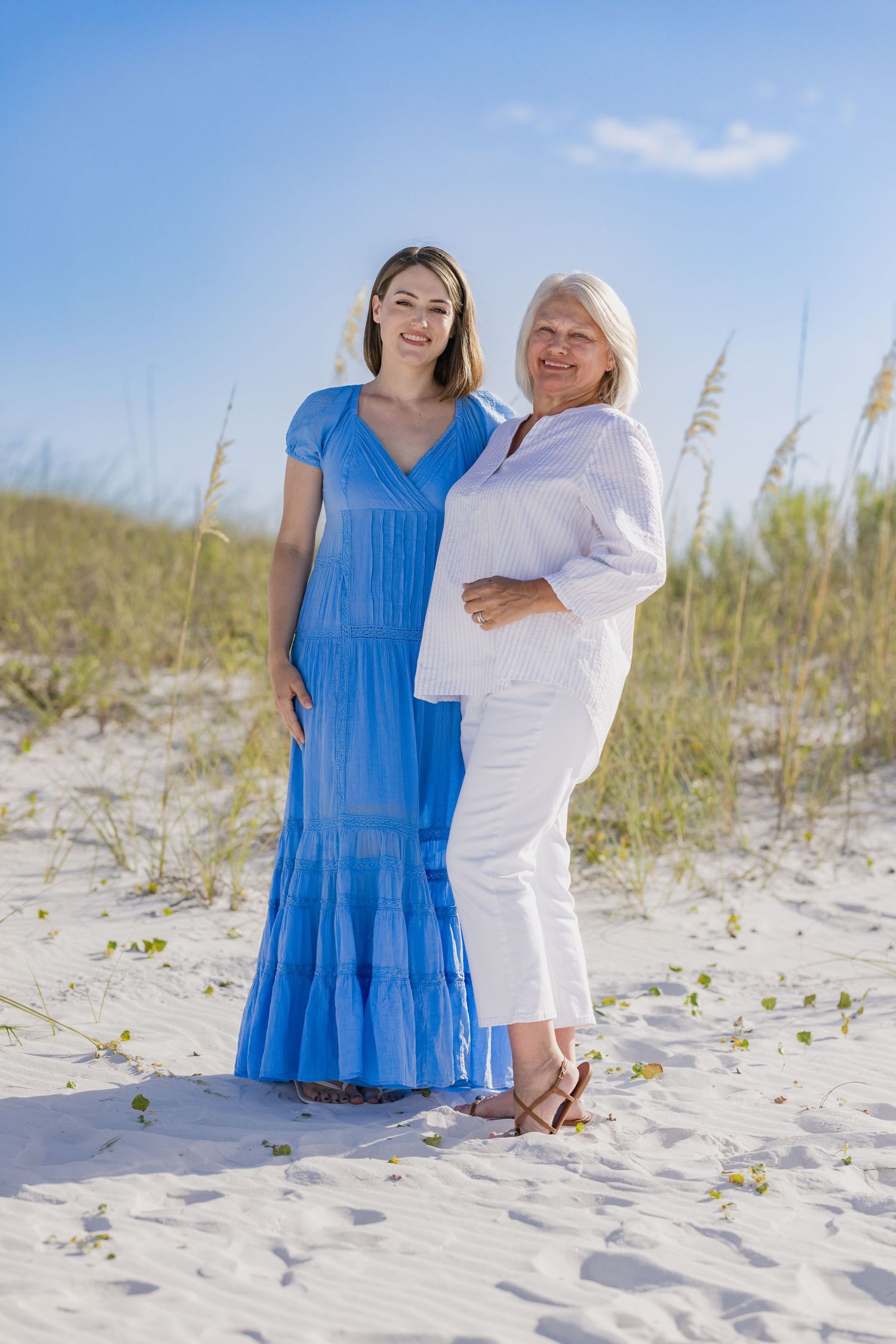 Two women, one in blue dress and one in white outfit, smiling together on a sandy beach with sea oats.