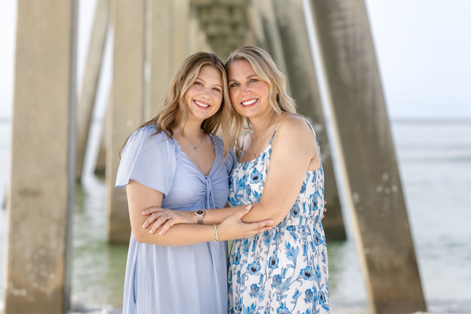 Two smiling women in blue dresses pose together under a beach pier on a sunny day.