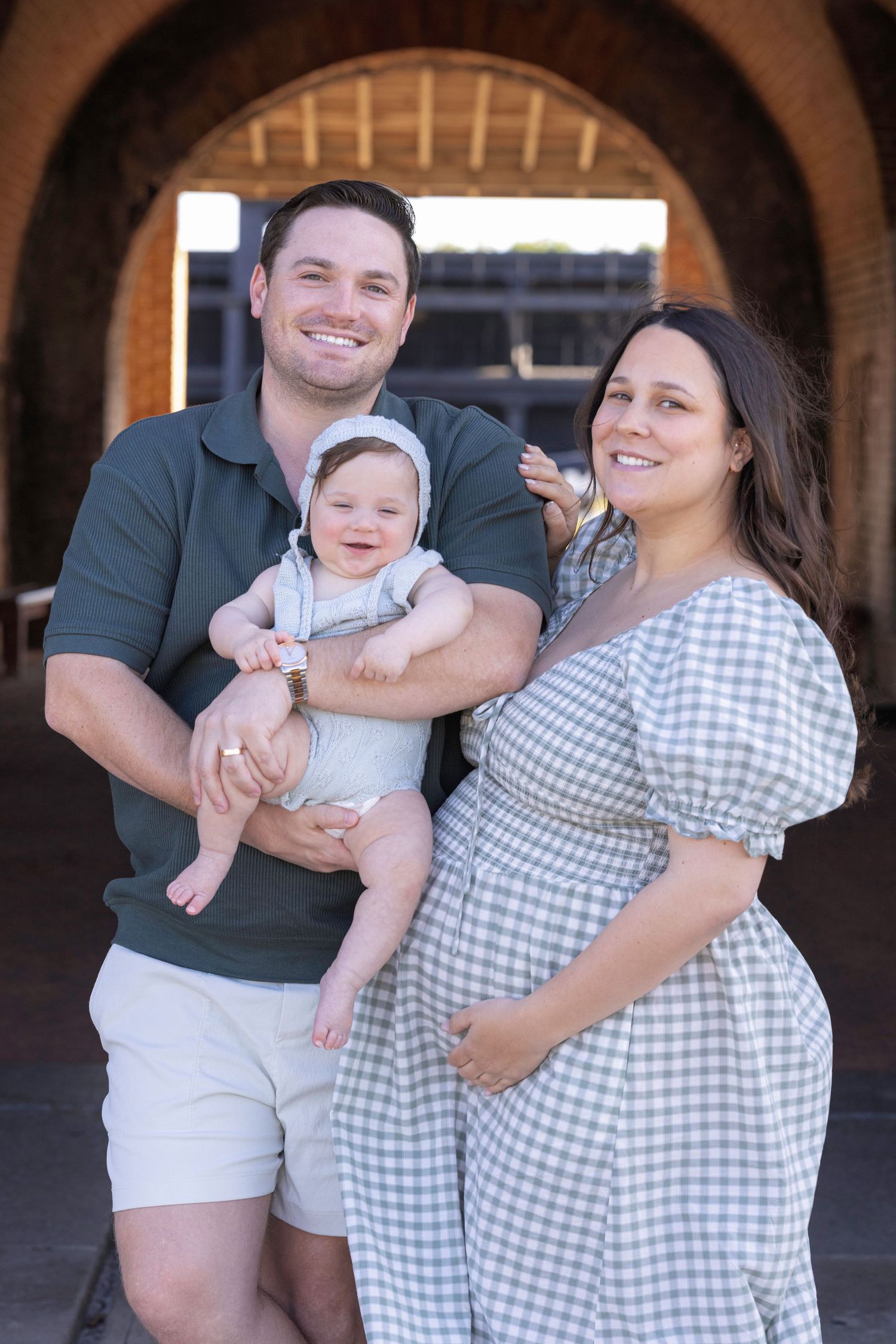 A smiling family with a man holding a baby in a bonnet beside a pregnant woman in a gingham dress under brick arches.