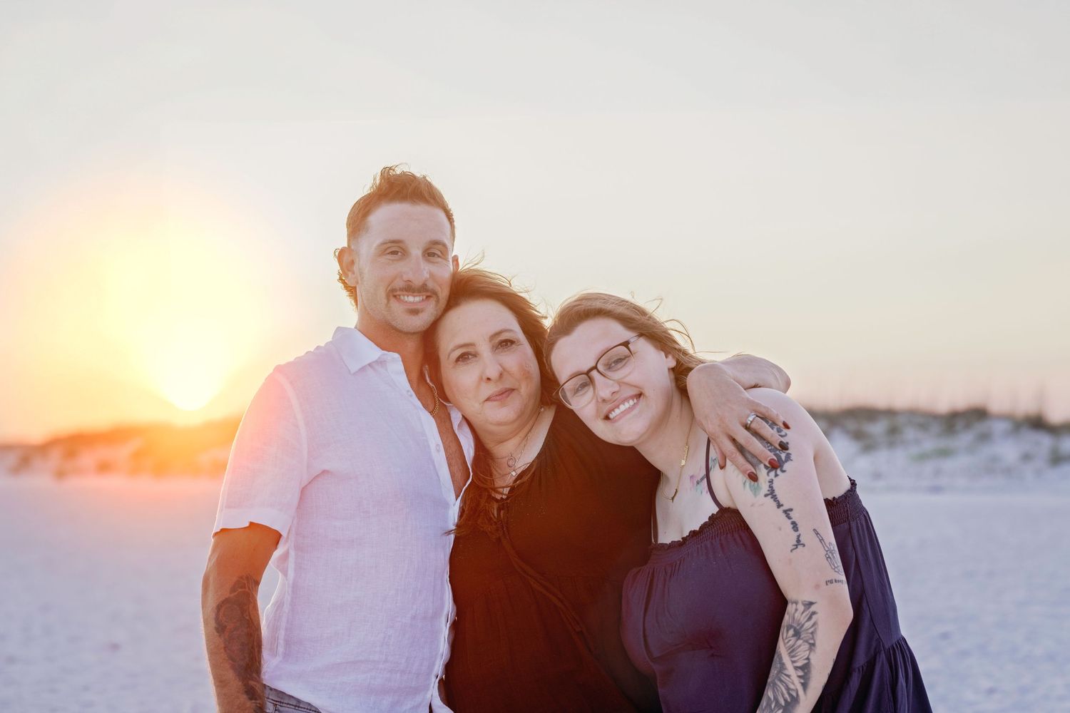 Three adults smiling together on beach at sunset in casual attire.