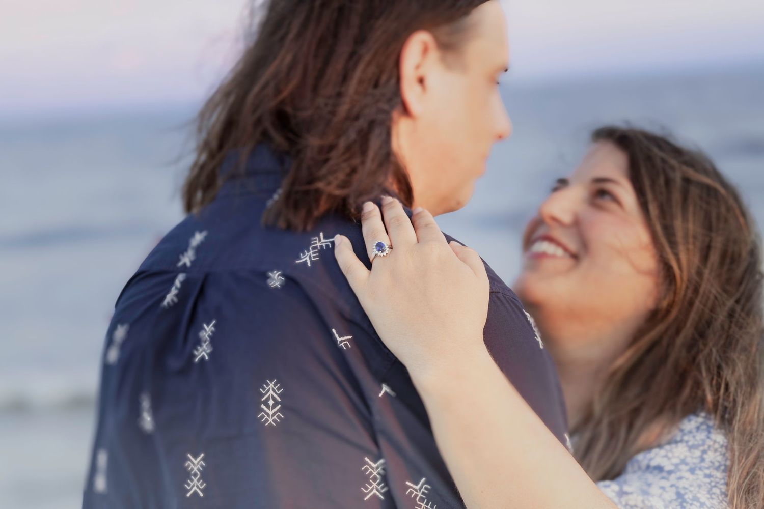 Couple embracing on a beach at sunset, woman smiling and showing engagement ring on her hand.