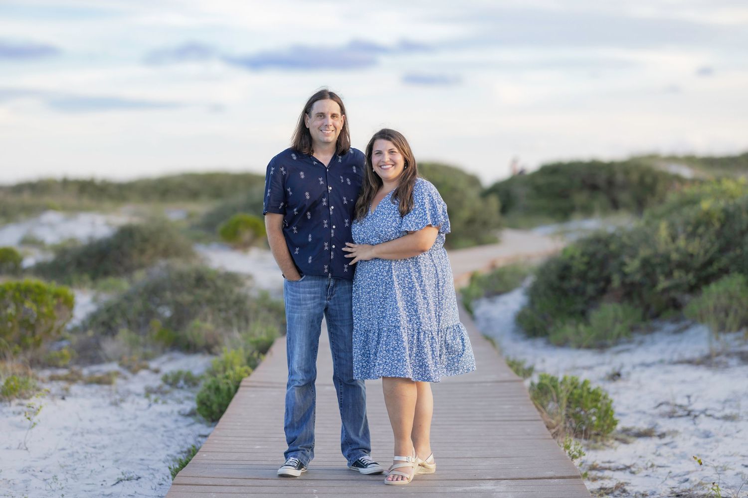 Couple posing on a wooden boardwalk path through coastal sand dunes at a beach during golden hour.