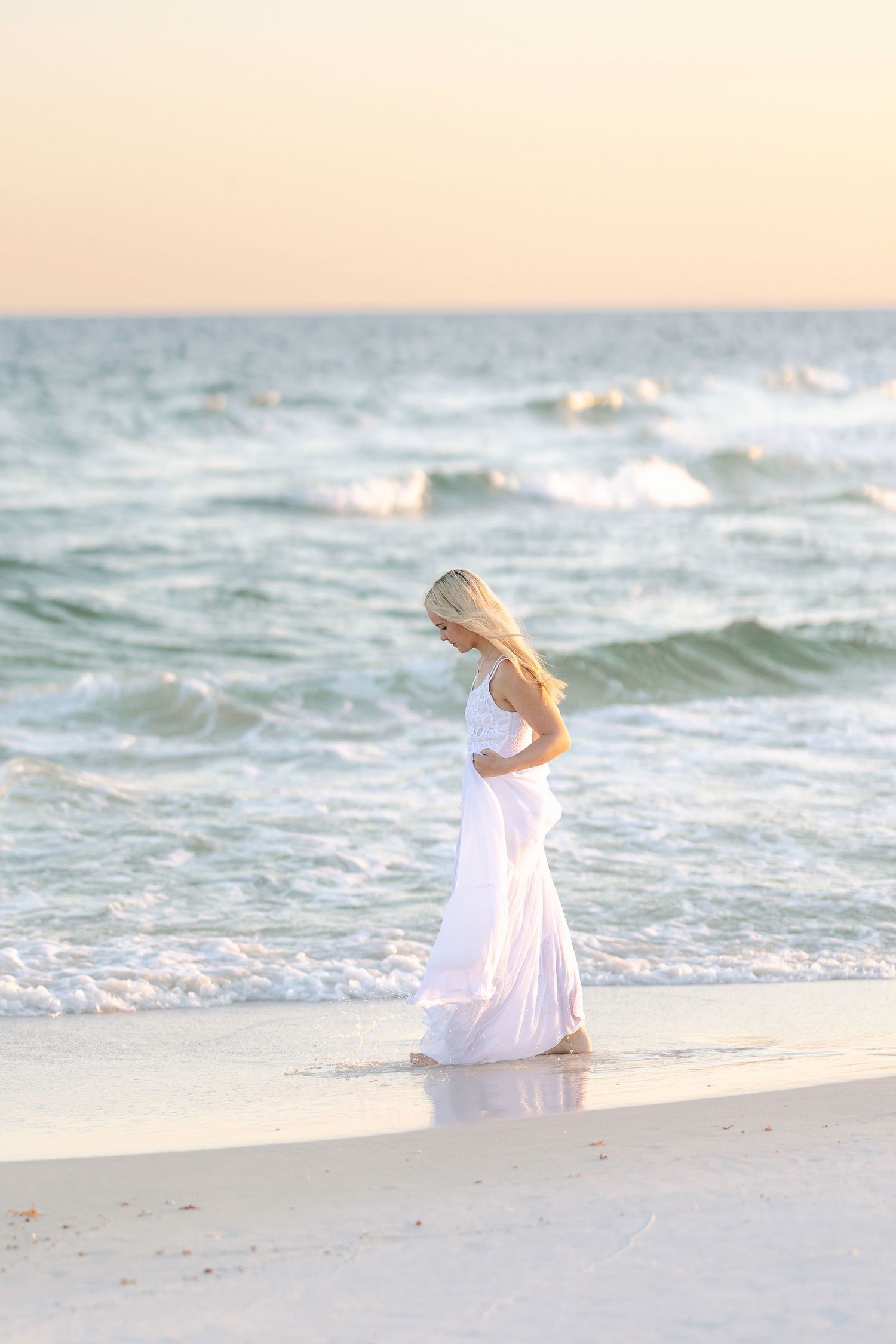 Blonde woman in white dress walking on a sandy beach at sunset with ocean waves in the background.